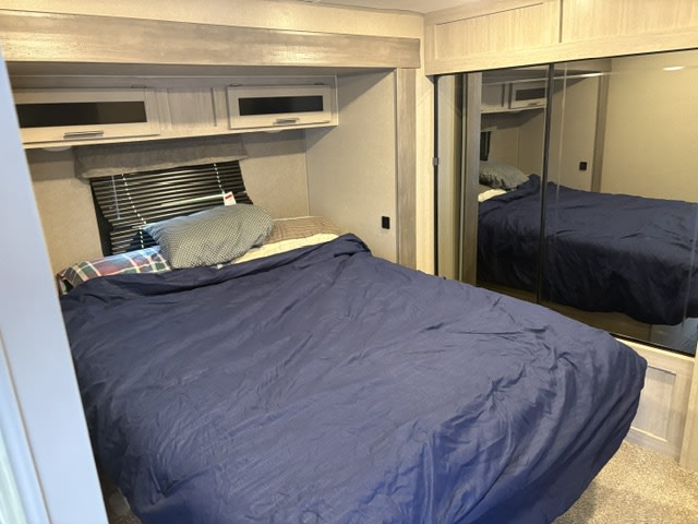 Spacious RV rental bedroom featuring a cozy bed, overhead cabinets, a window with blinds, and a mirrored wardrobe.. Coachmen Catalina Legacy 2022