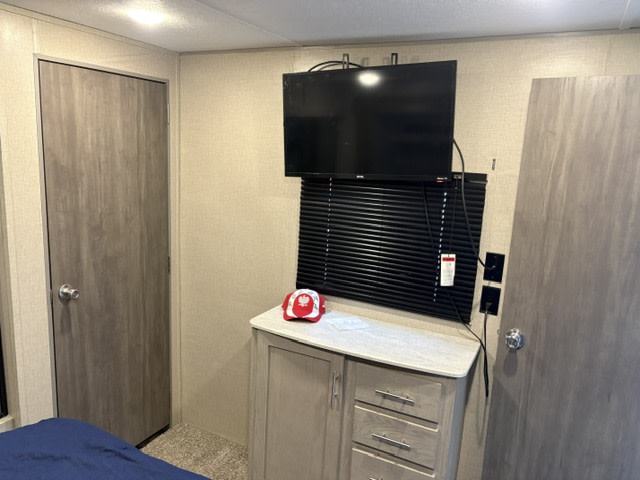 This RV rental bedroom features a wall-mounted TV, black window blinds, a storage cabinet with a red cap, and two light wood doors.. Coachmen Catalina Legacy 2022