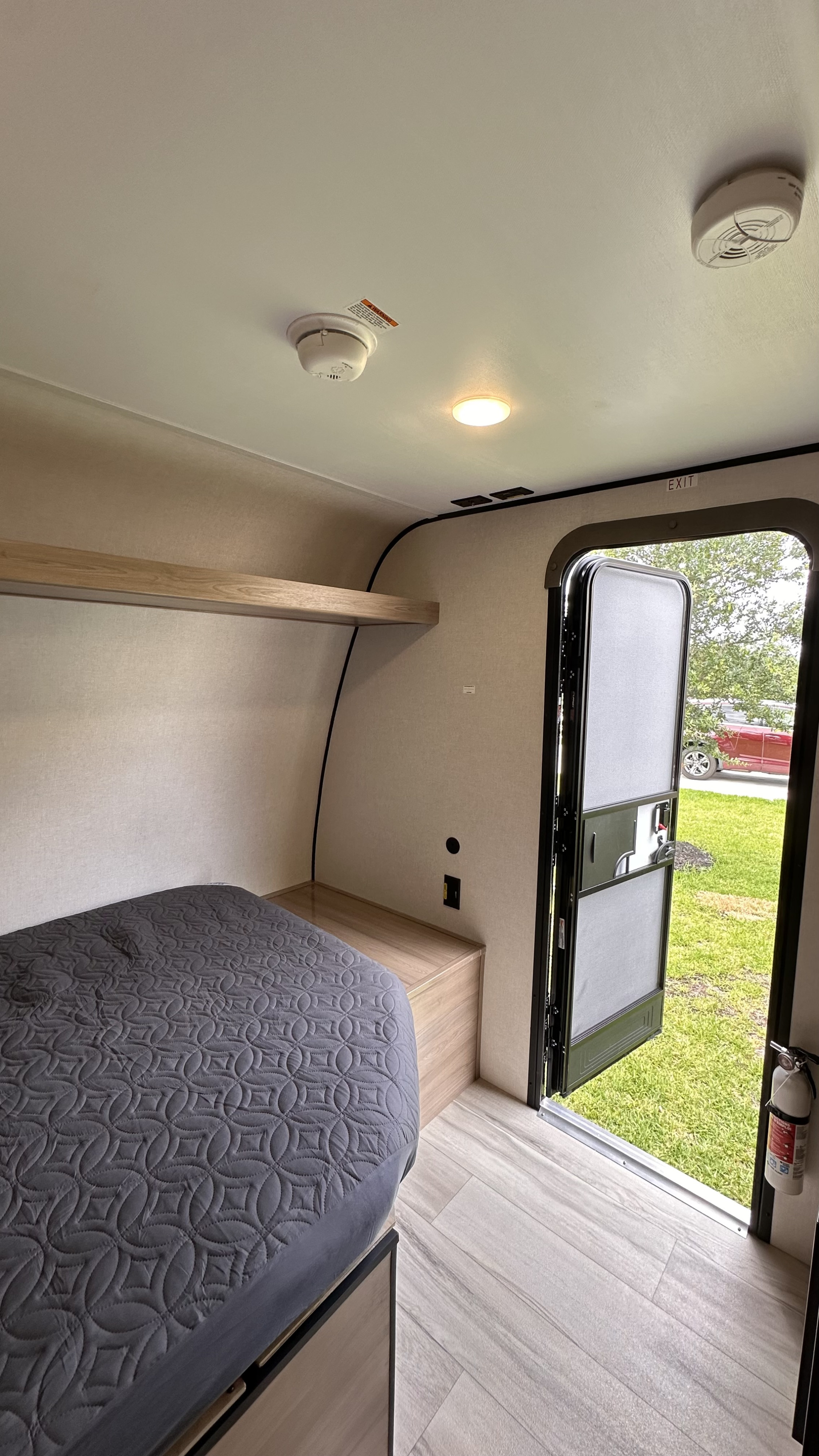 RV rental interior: cozy bed, wooden shelf, smoke alarms, fire extinguisher by the open door to the outdoors.. Keystone RV 13B travel Trailer 2026