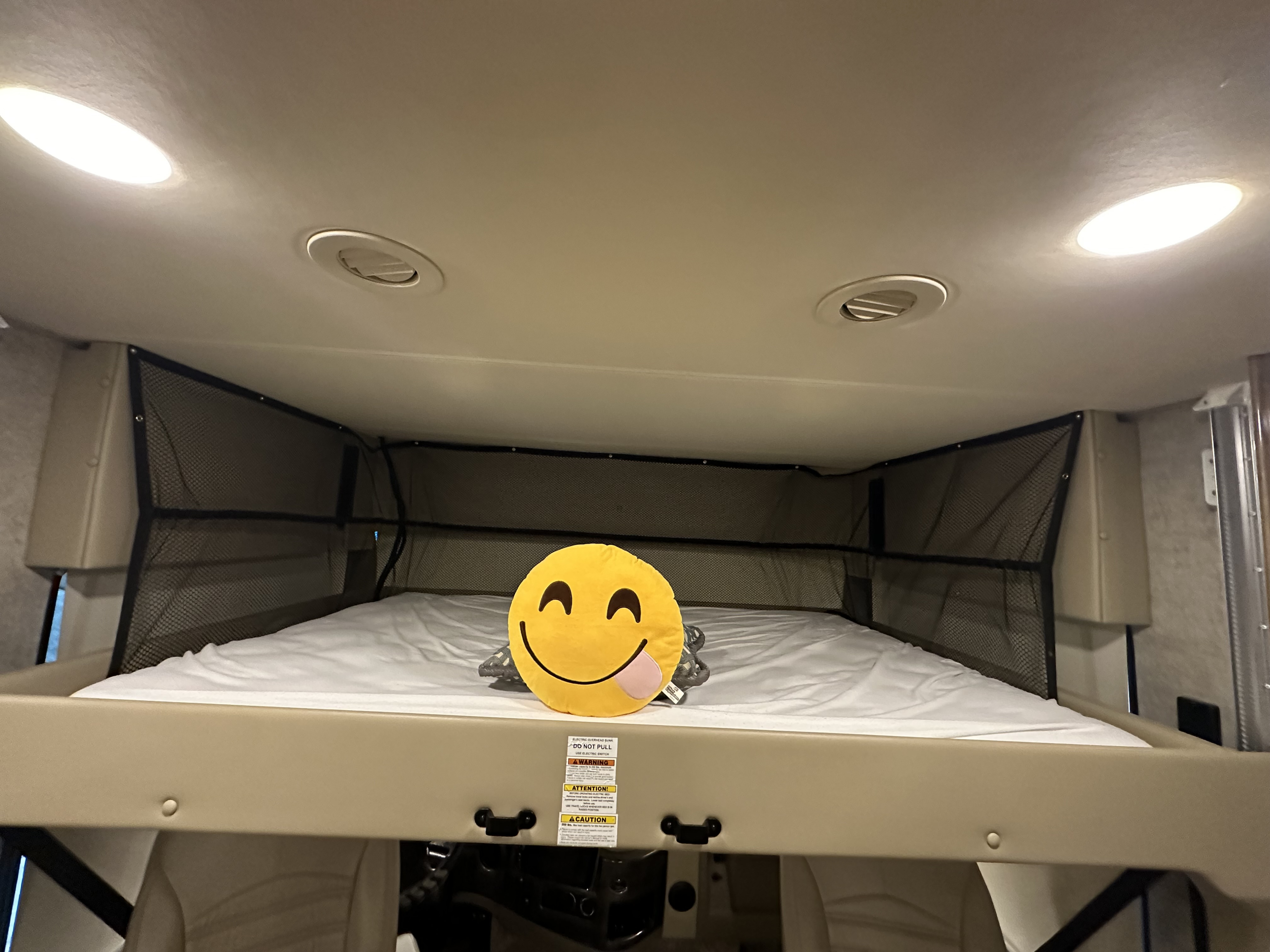 Cozy RV rental overhead bunk with a comfy bed, safety netting, a cheerful smiley pillow, lights, and vents.. Thor Miramar 2020