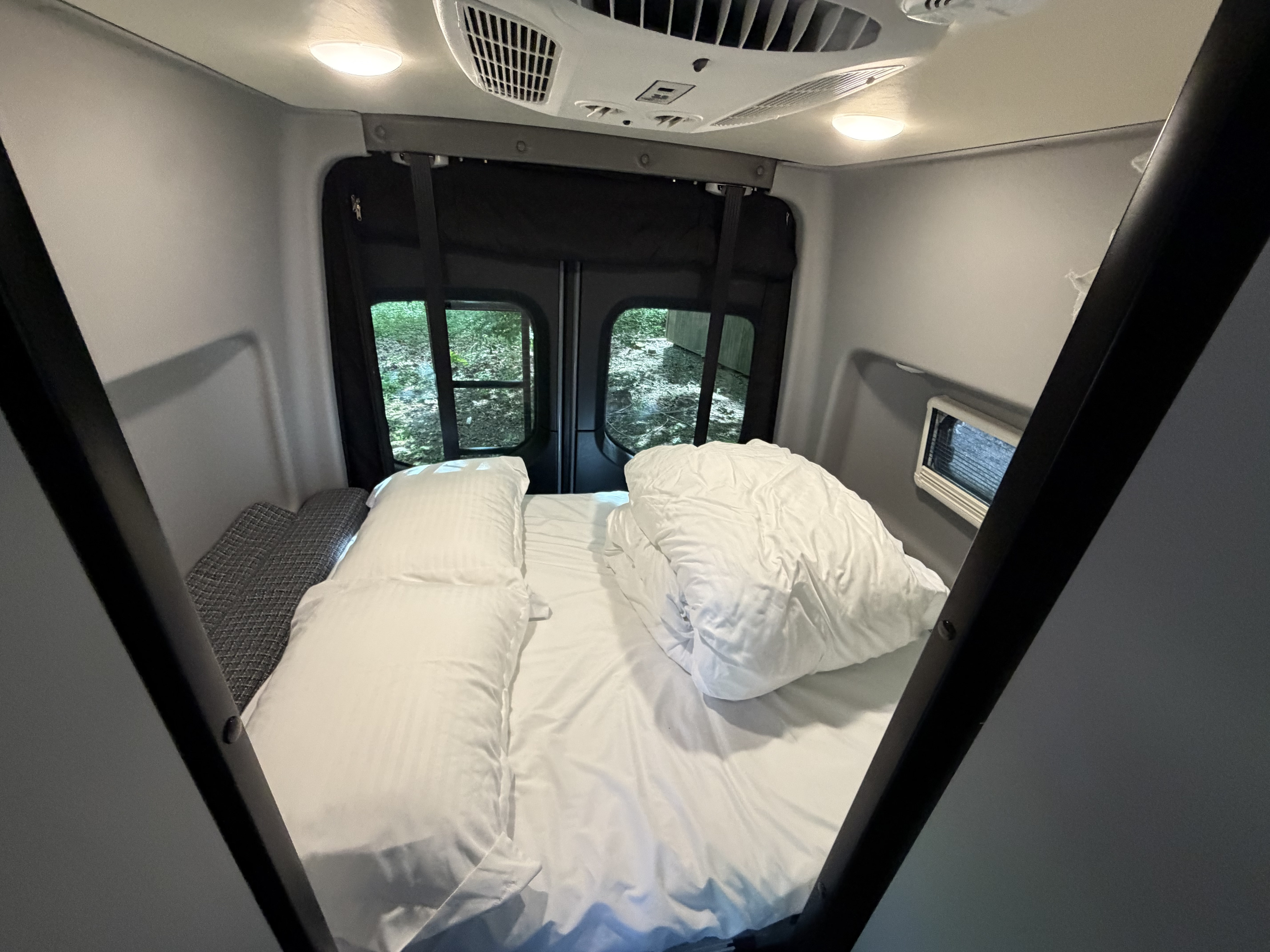 Step inside this cozy RV rental's sleeping area! Features a comfortable bed with white linens and rear window views of nature.. Winnebago Revel 2022