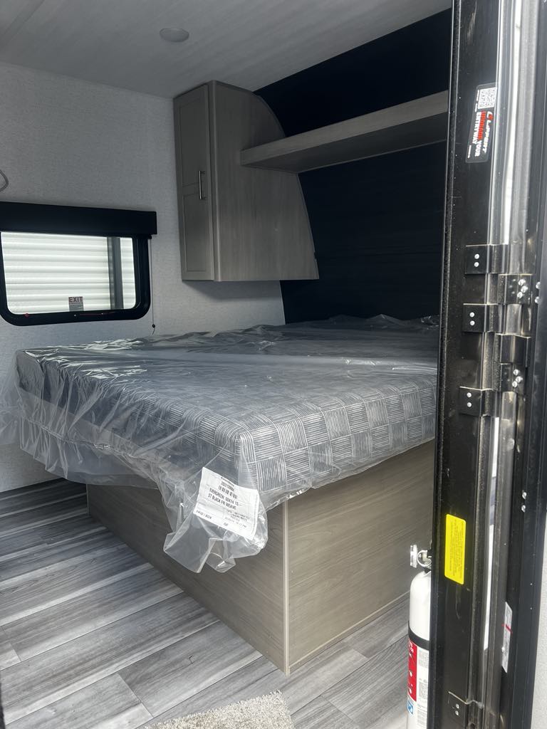 Clean RV rental bedroom with new bed, plastic-covered mattress, overhead storage, and window. Perfect for your next trip.. Keystone RV Springdale 2024