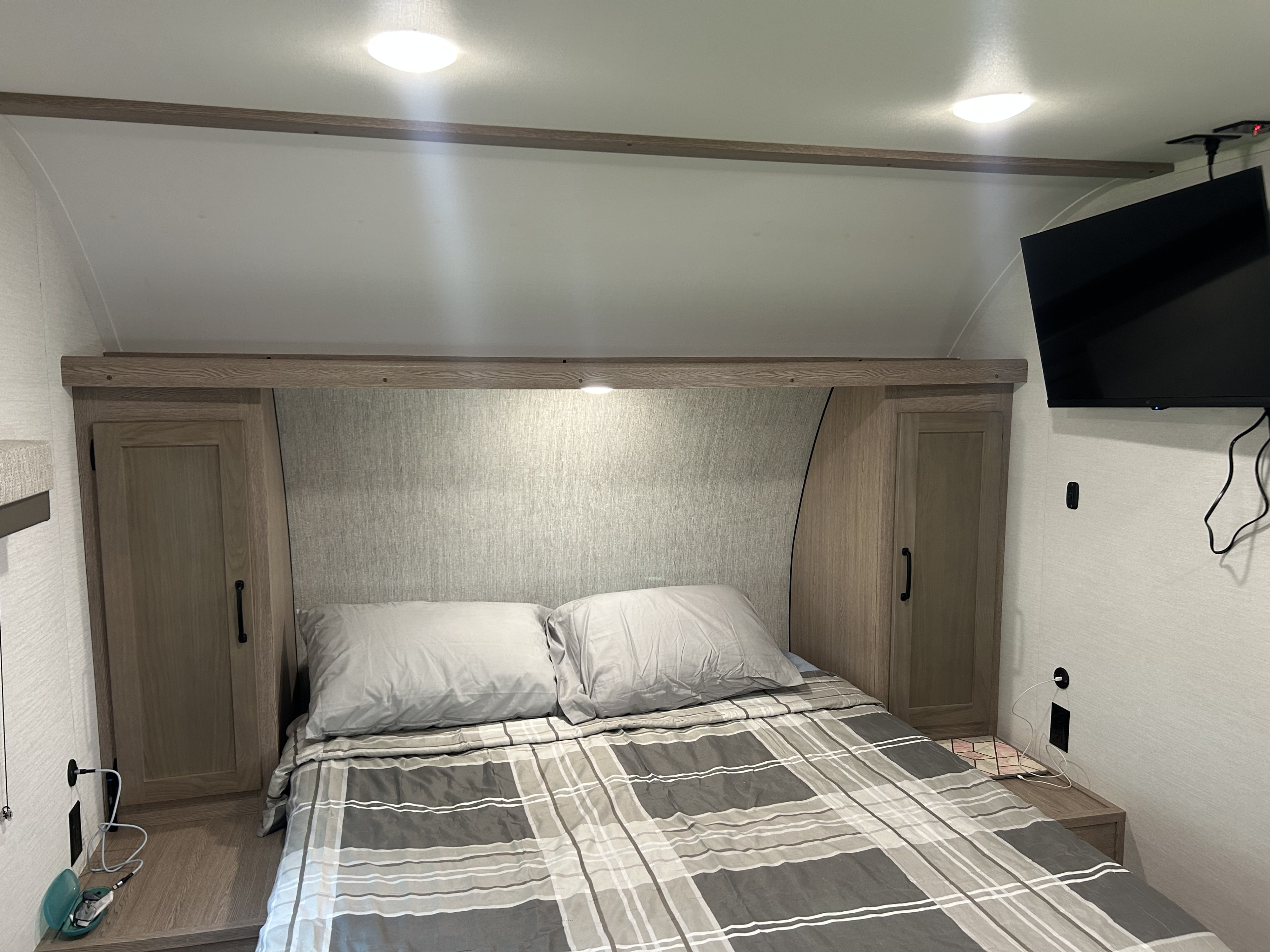 This RV rental shows a compact, modern bedroom with a plaid bed, side cabinets, overhead lights, and a TV.. Gulf Stream Grand River 25BH 2026