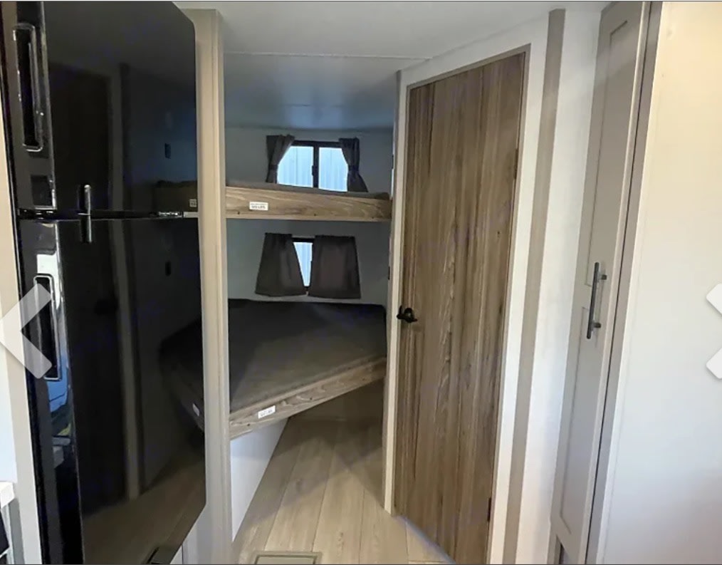 This RV rental interior shows a black refrigerator left, a bunkhouse with two beds ahead, and a wooden door to the right.. KZ Connect SE 2026