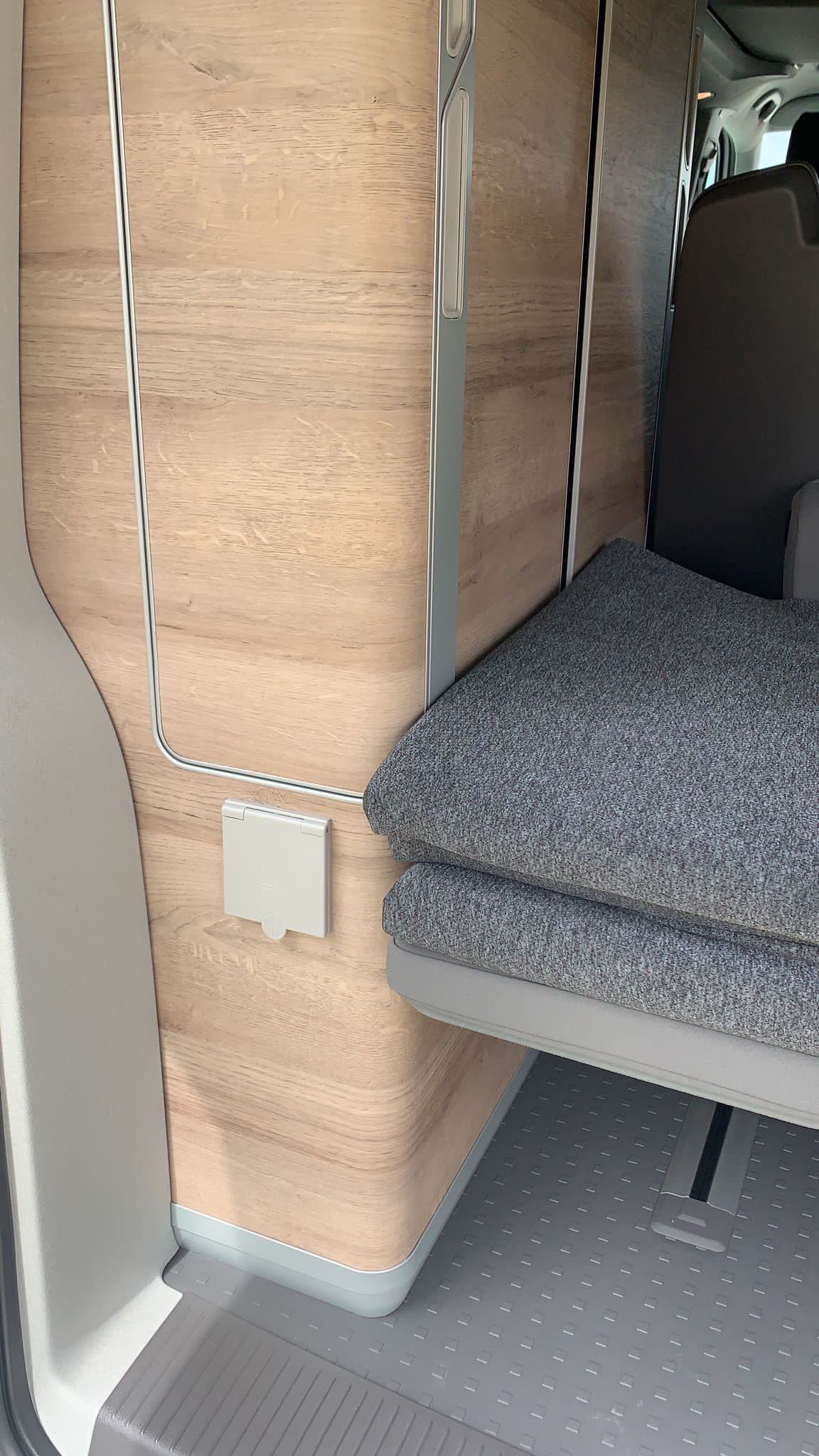 RV rental interior featuring wood cabinetry, stacked grey cushions, a wall power outlet, and textured rubber flooring.. VOLKSWAGEN CALIFORNIA 2023