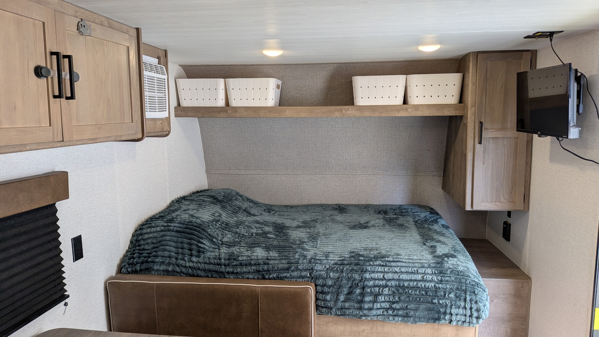 Cozy RV rental bedroom features a plush blue blanket on the bed, overhead storage baskets, light wood cabinets, and a wall-mounted TV.. Keystone RV Springdale Mini 2025