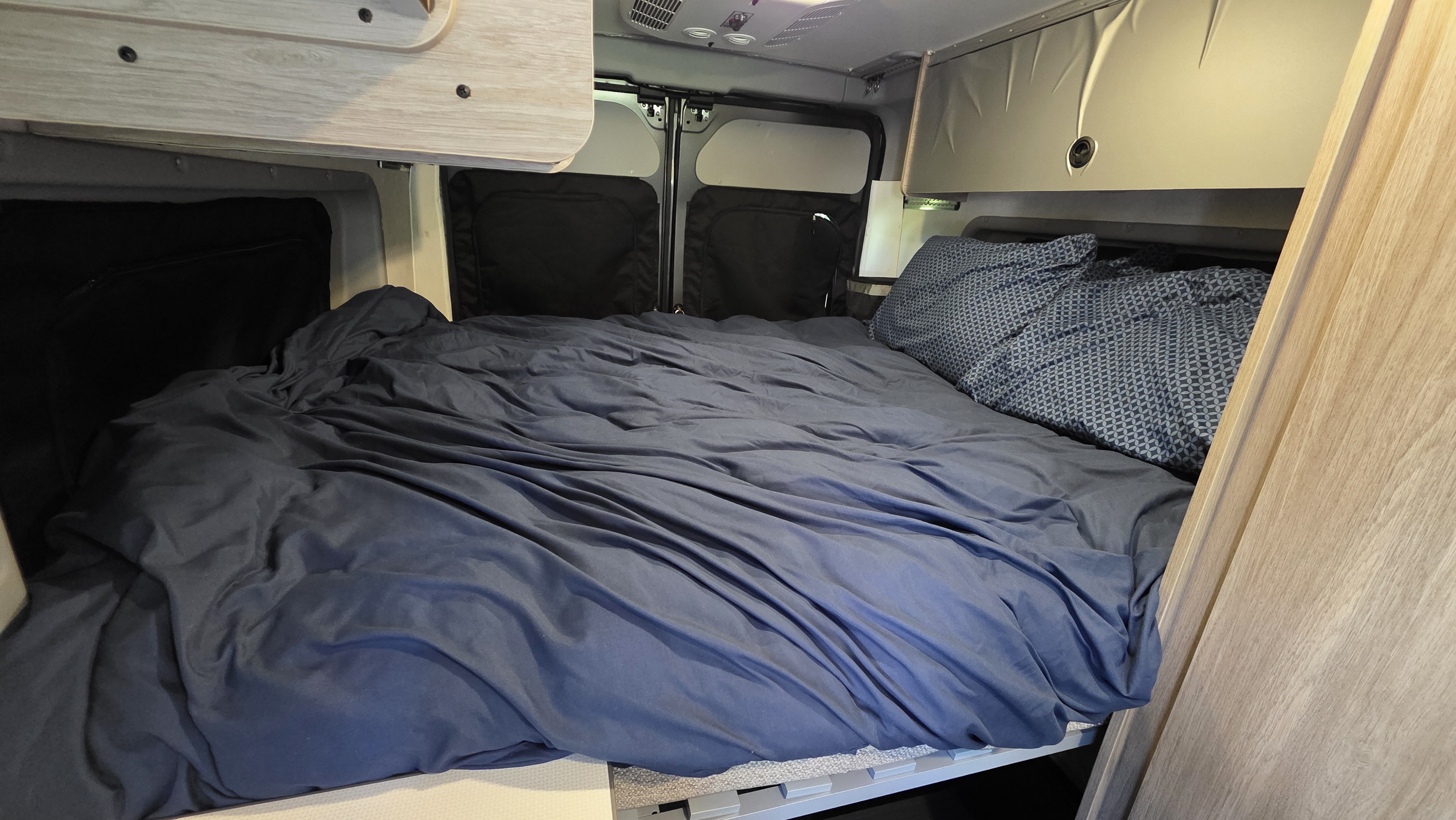 Cozy bed inside this RV rental, featuring dark blue sheets and a patterned pillow, offering a comfortable sleeping area for your travels.. Winnebago Solis 2023