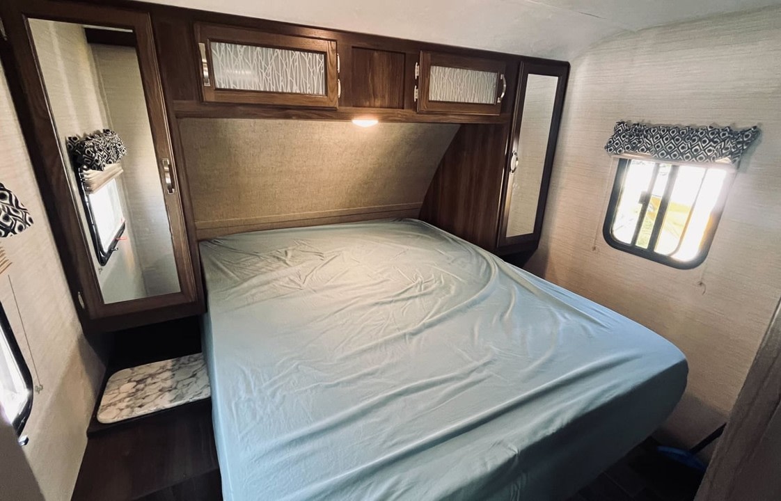 Cozy RV rental bedroom featuring a bed with light blue sheets, overhead storage, mirrored cabinets, and side windows.. Keystone RV Bullet Ultra Lite 2019