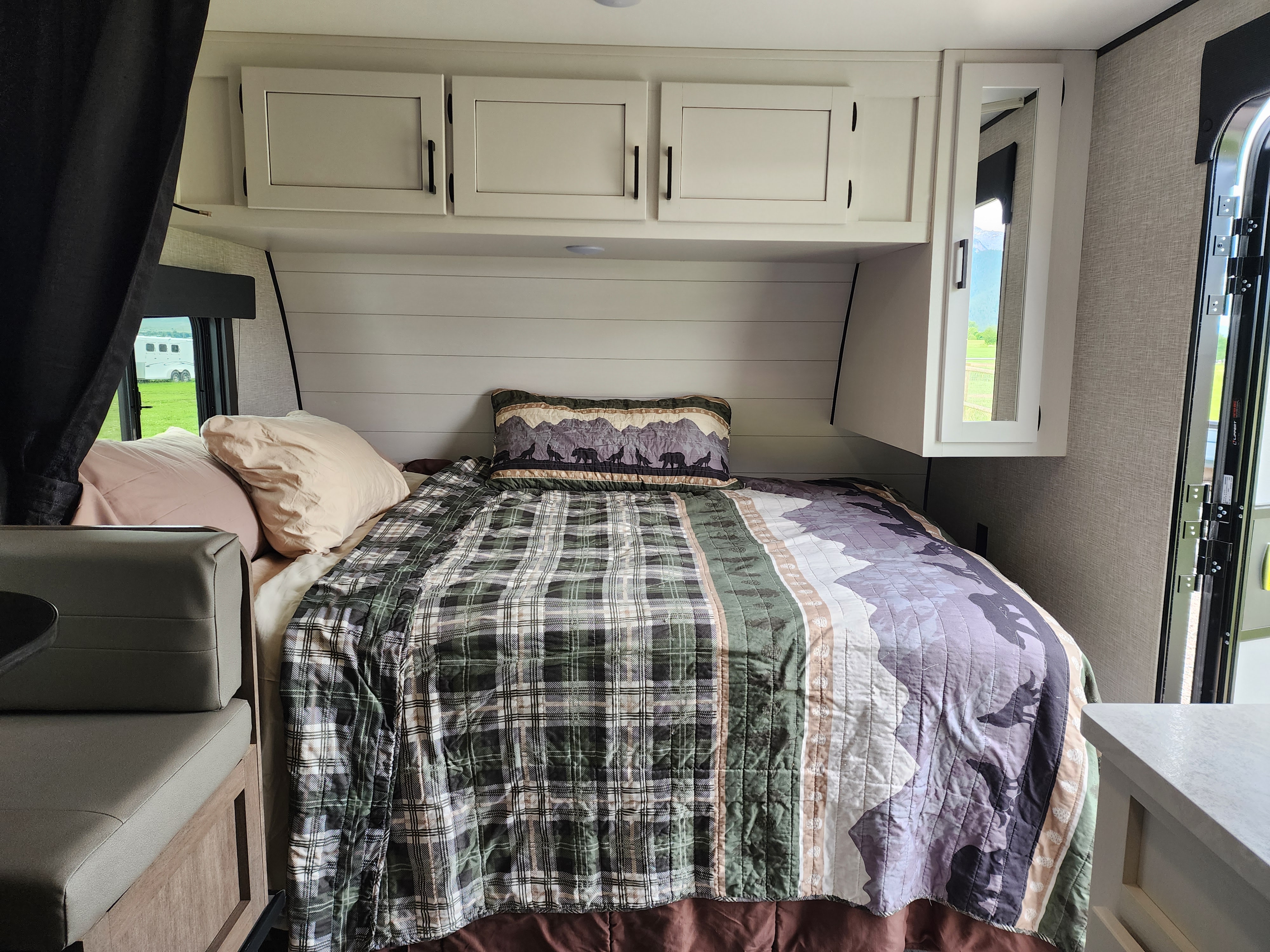 Cozy RV rental bedroom with plaid and mountain bedding, overhead cabinets, and a scenic window view.. Highland Ridge RV Open Range 2024