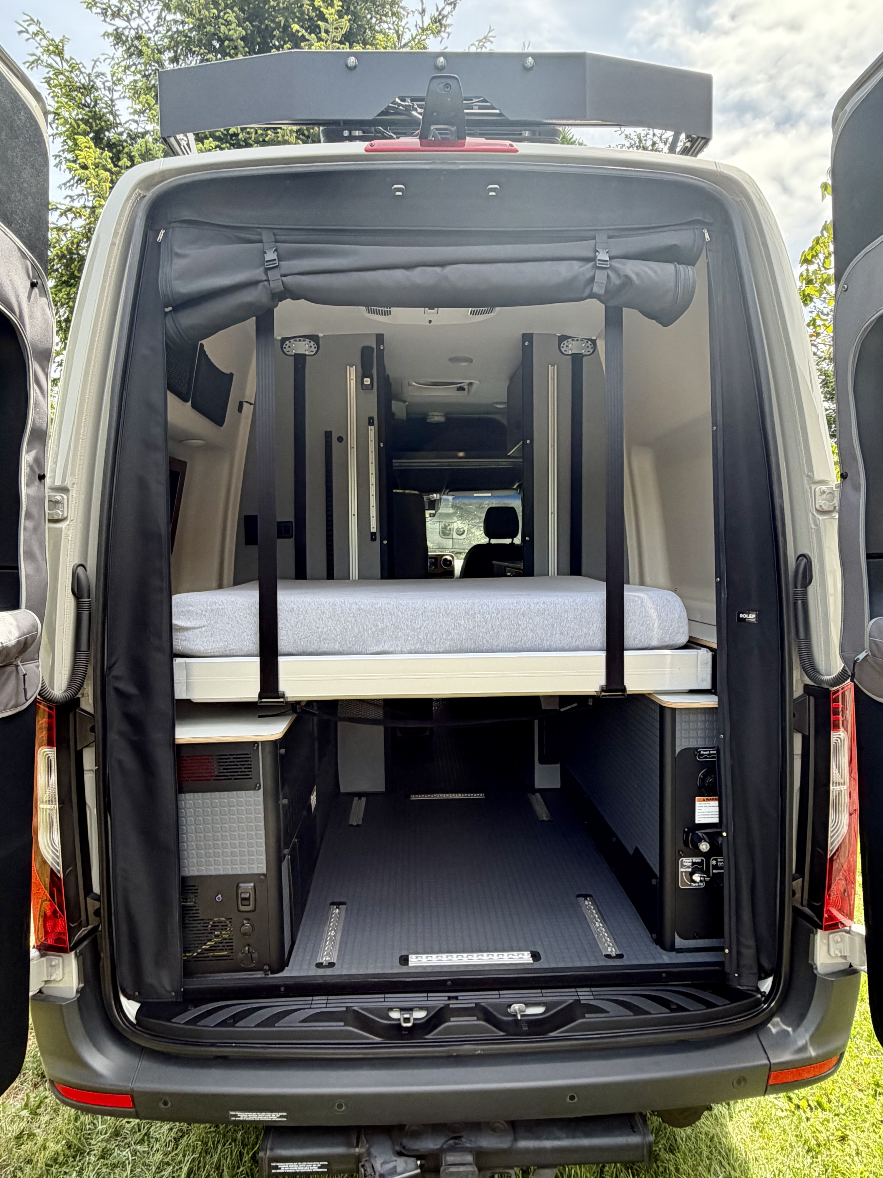 Inside a spacious RV rental camper van, rear doors open to reveal a comfortable elevated bed and functional storage.. Winnebago Revel 2025