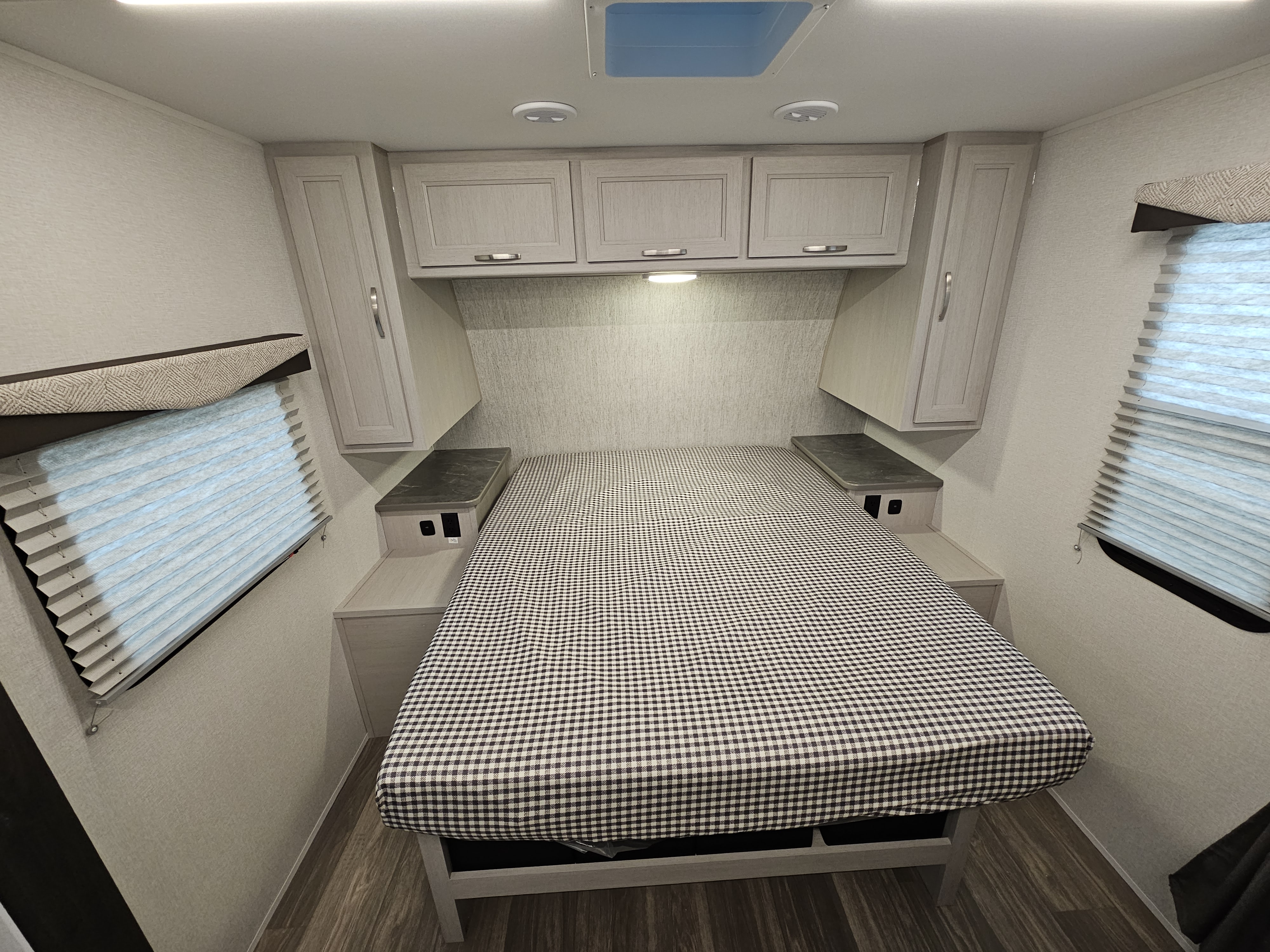 Cozy RV rental bedroom with a checkered comforter on the bed, overhead cabinets, side tables, and windows.. Cruiser RV Shadow Cruiser 2025