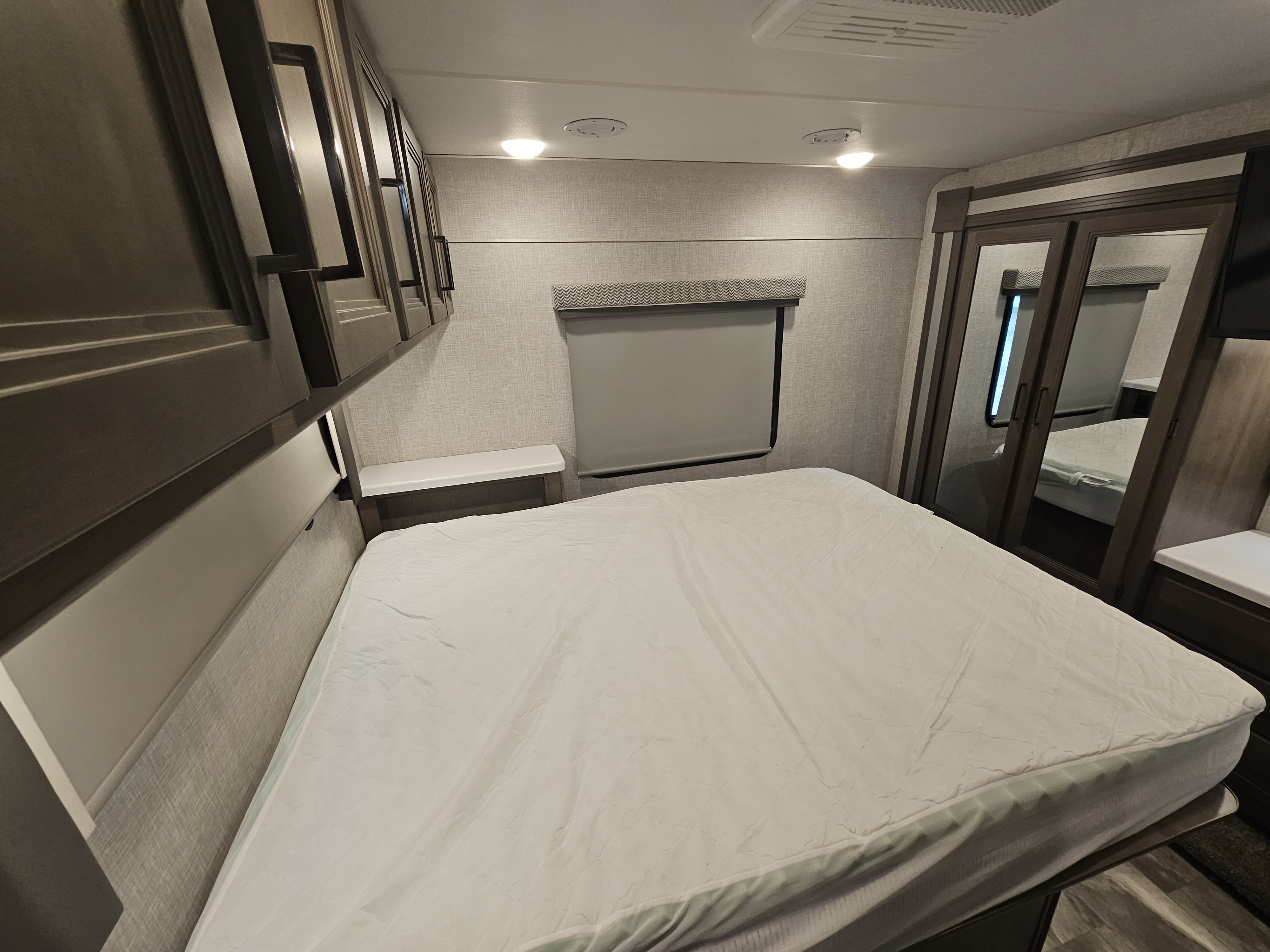 Cozy RV rental bedroom features a comfy bed, overhead cabinets, window blind, and mirrored closet for travel comfort.. Thor Quantum 2023