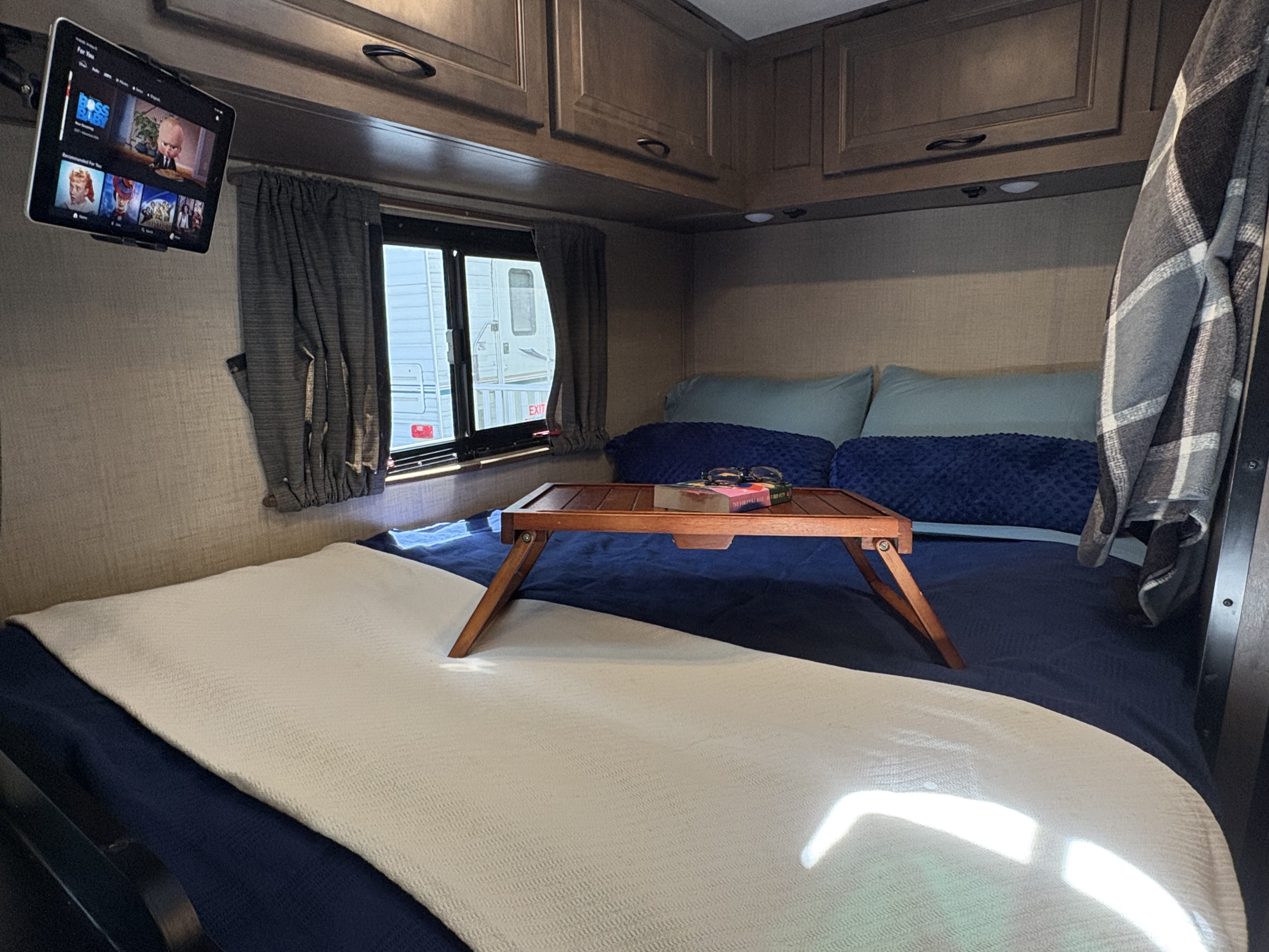 Cozy RV rental bedroom featuring a bed with a wooden tray, tablet with movies, and a window view of another RV.. Tiffin Adventure 2020
