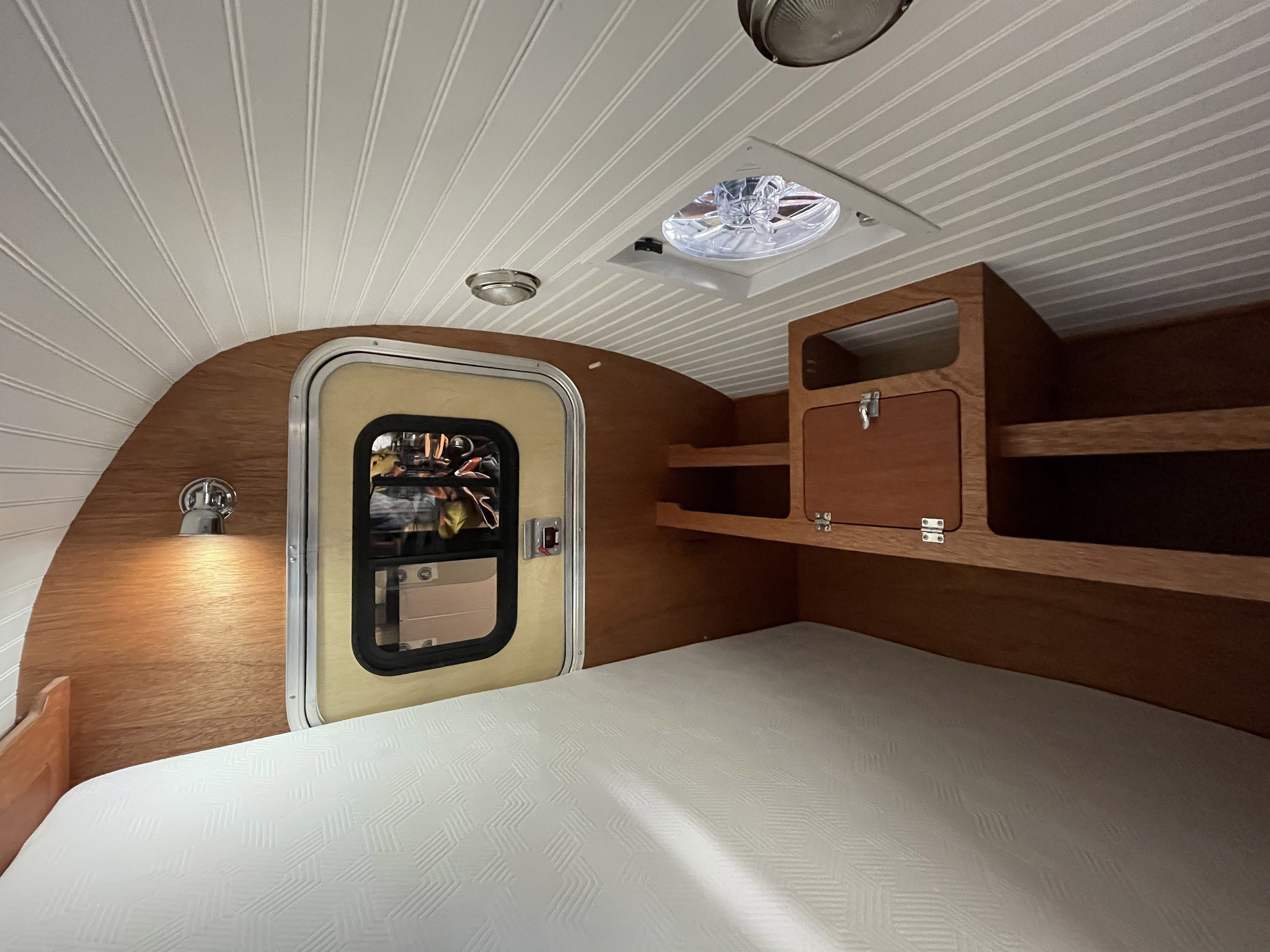 Interior of an rv rental featuring a clean bed, wood walls, built-in storage, a door, and overhead fan/lights.. All Good Equipment Company Teardrop Trailer 2026