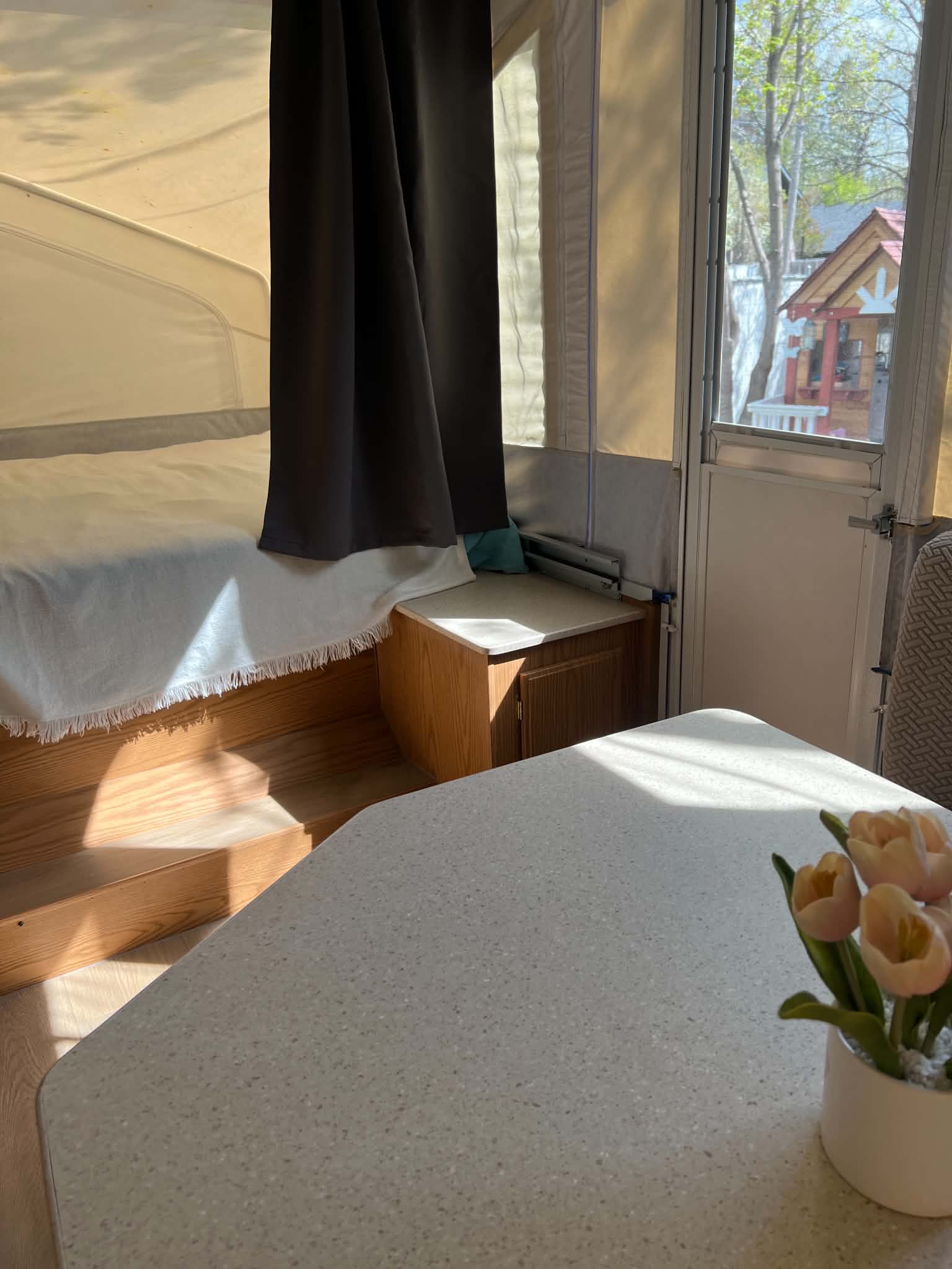 Cozy rv rental interior: bright bed, dinette table with tulips, and a sunny outdoor view of trees & a small playhouse.. Flagstaff Towing 2005