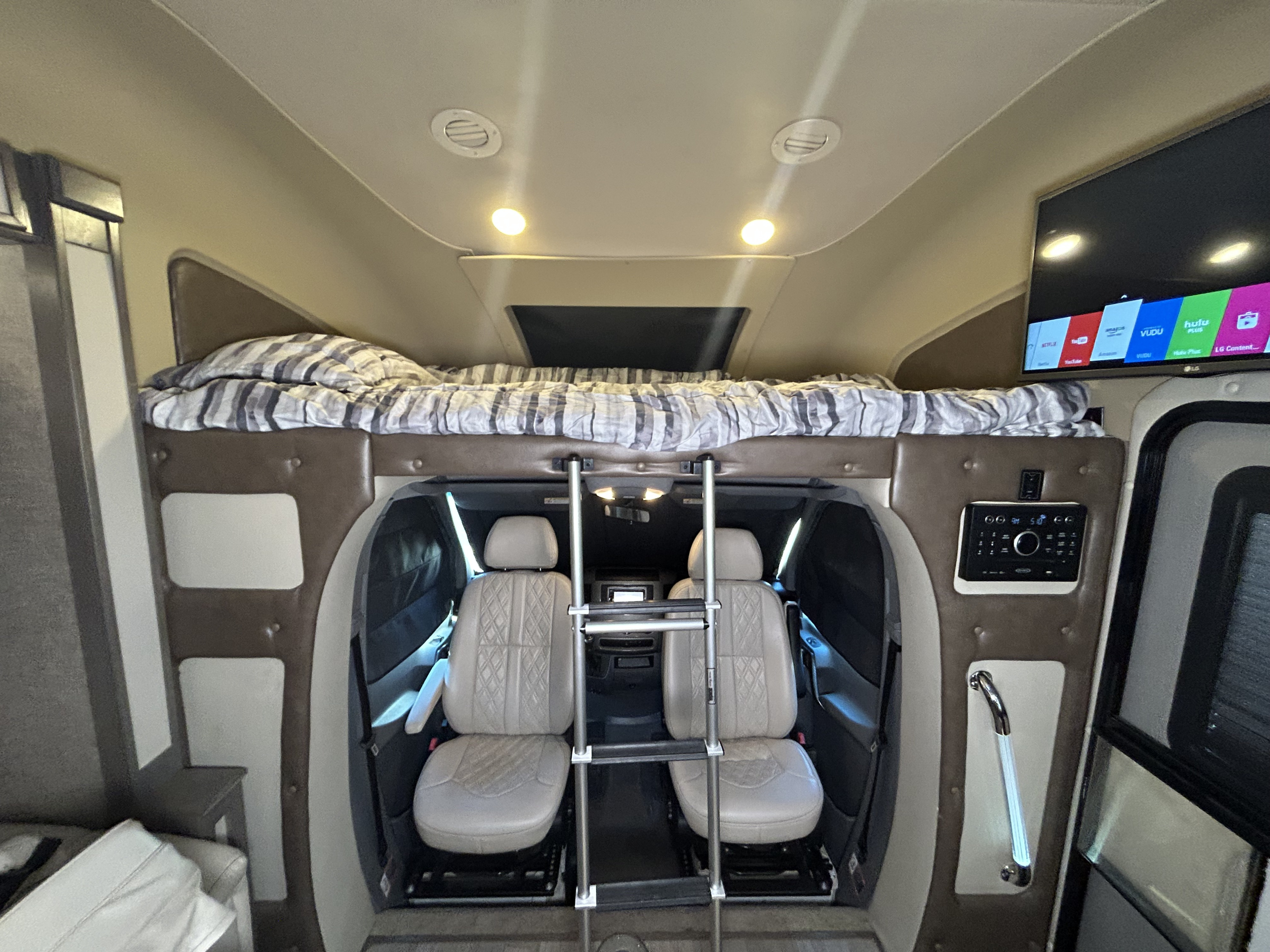 Cozy RV rental interior with an elevated sleeping bunk (striped bedding), a ladder, swivel front seats below, and a TV displaying apps.. Tiffin Wayfarer 2017