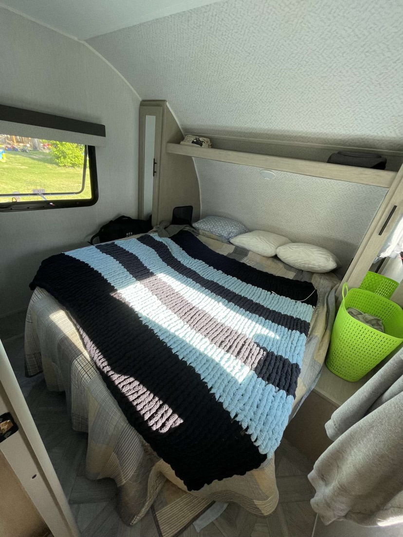 Cozy RV rental bedroom featuring a bed with a striped blanket, window view, and overhead storage for a comfortable travel experience.. Forest River Wildwood X-Lite 2023