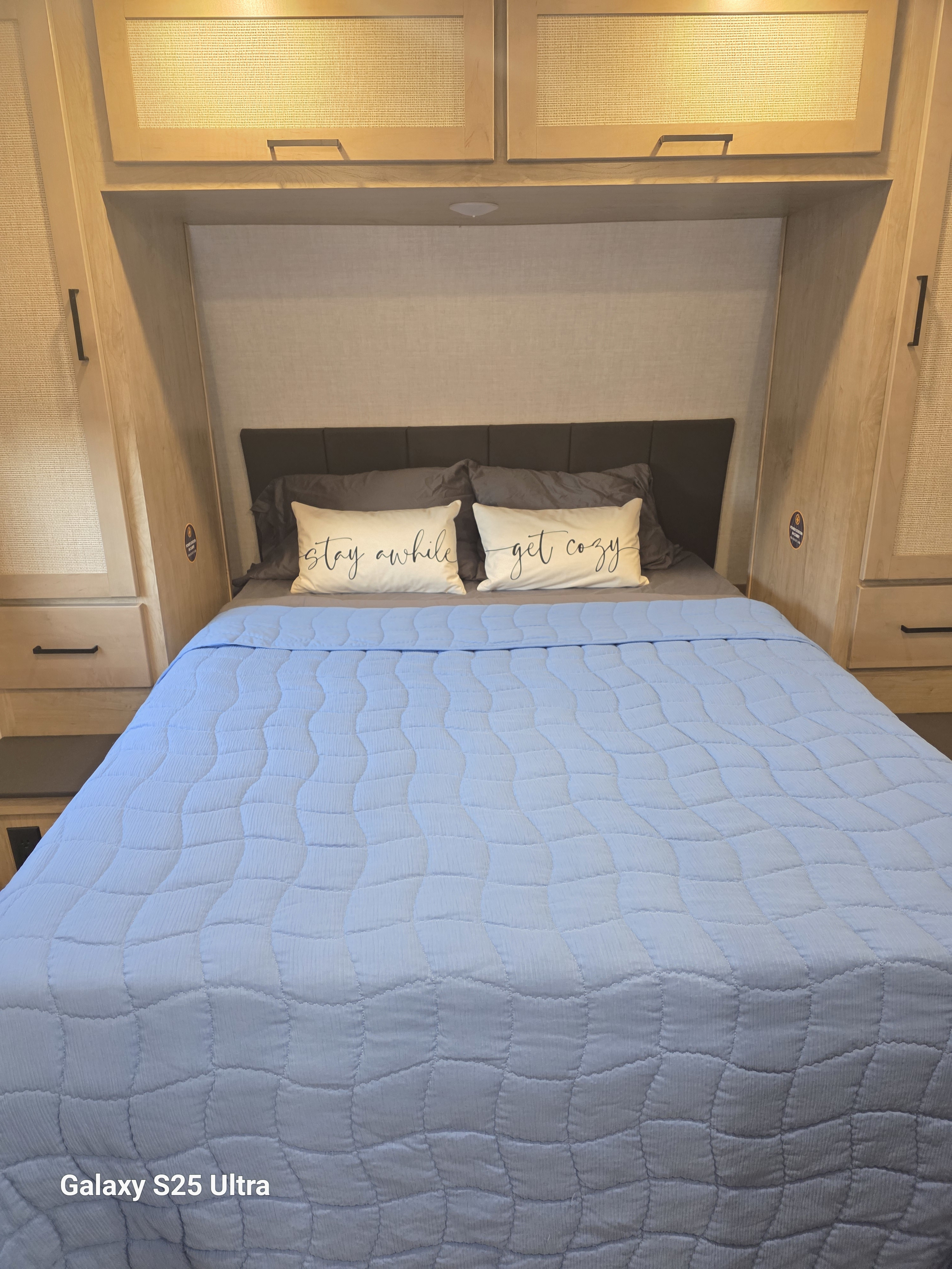 Cozy RV rental bedroom featuring a light blue quilted bed, "stay awhile get cozy" pillows, and warm wood cabinets.. Ember E-Series 2024