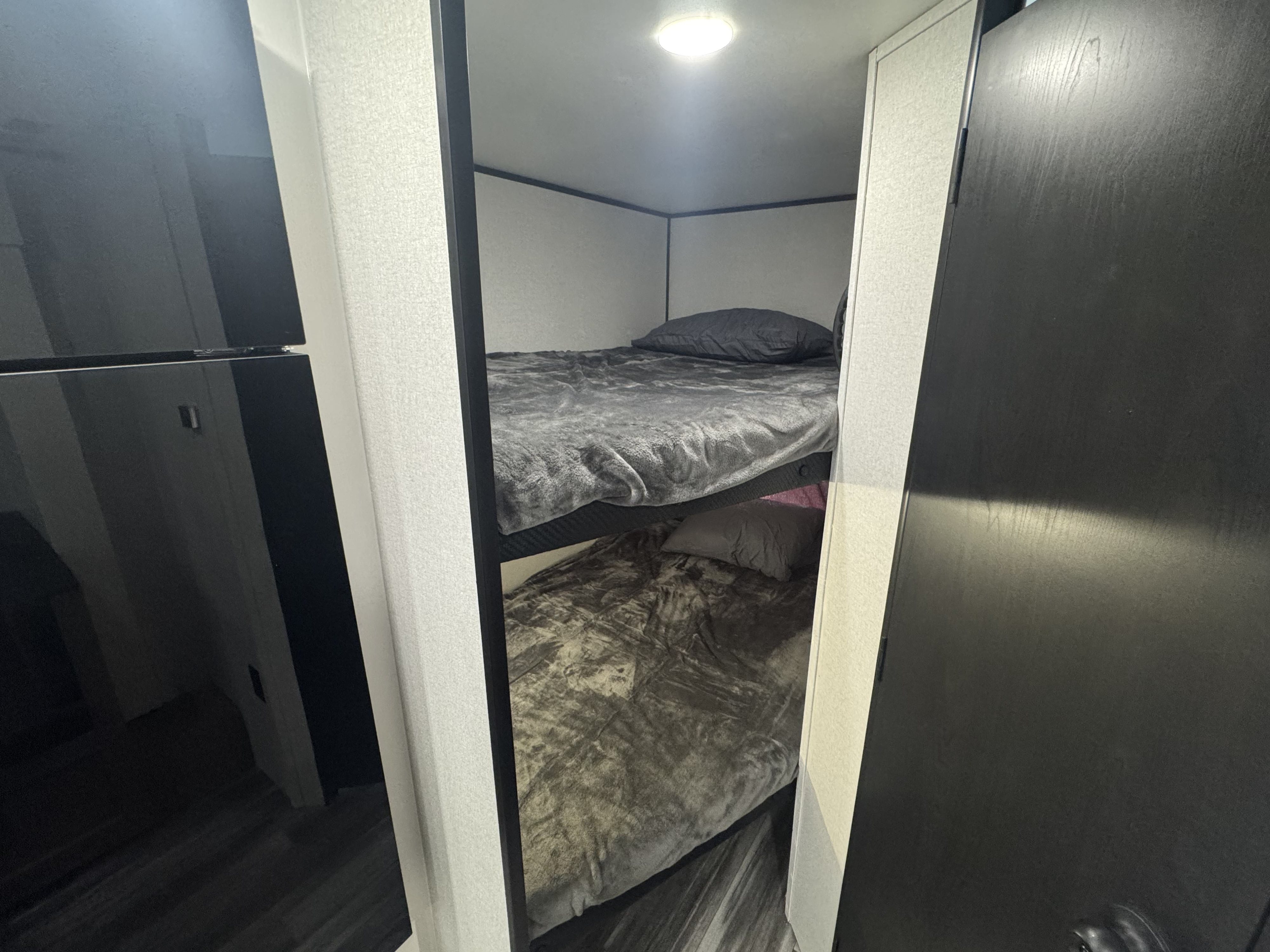 RV rental bunk beds with gray bedding and a ceiling light. Compact sleeping area shown, with a reflective fridge on the left.. Jayco Jay Flight SLX 197MBW Baja 2026