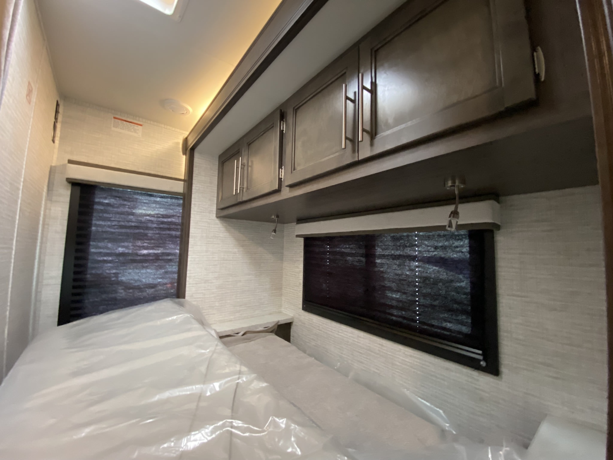 Clean RV rental bedroom with new plastic-covered bed, dark overhead cabinets, and window blinds. Ready for adventure.. Entegra Coach Odyssey SE 2025