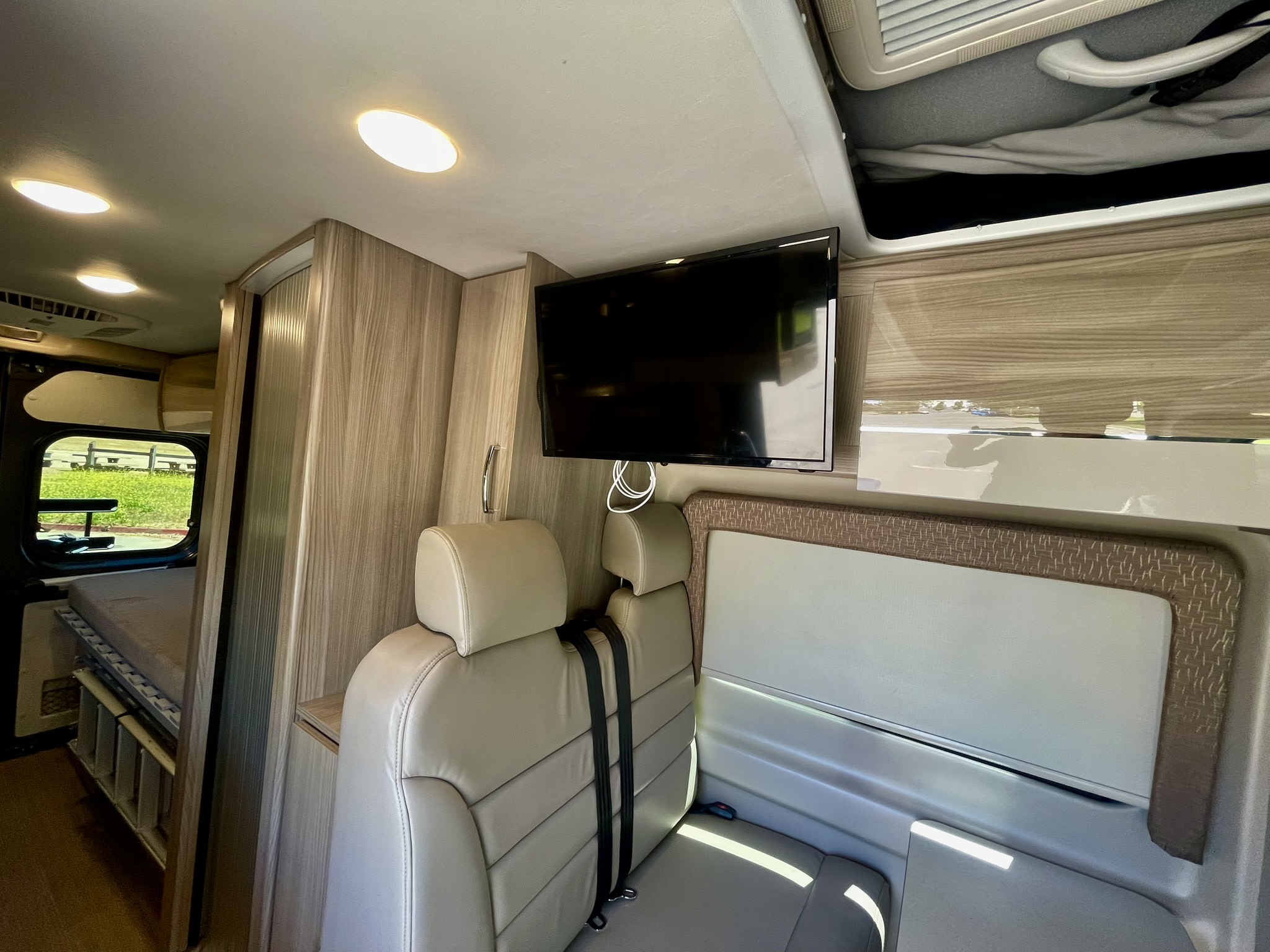 Interior of a modern RV rental, showcasing light seating, a mounted TV, overhead lights, wood cabinets, and a rear bed.. Thor Sequence 2022