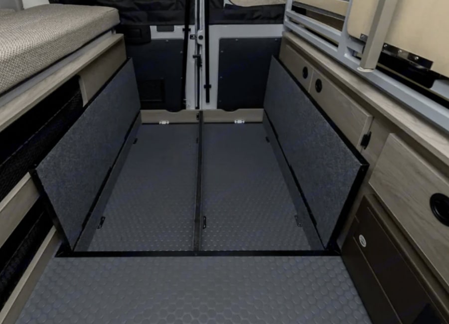 Rear view of an rv rental's interior, showing two deep, gray-lined storage compartments, a textured floor, and a bed above.. Winnebago Solis 2021