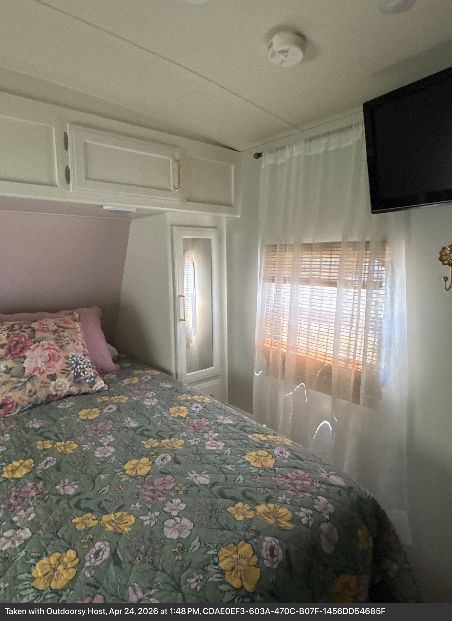 Bright rv rental bedroom featuring a floral bed, overhead cabinets, a window with curtains/blinds, and a wall-mounted TV.. Grand Design Imagine 2017