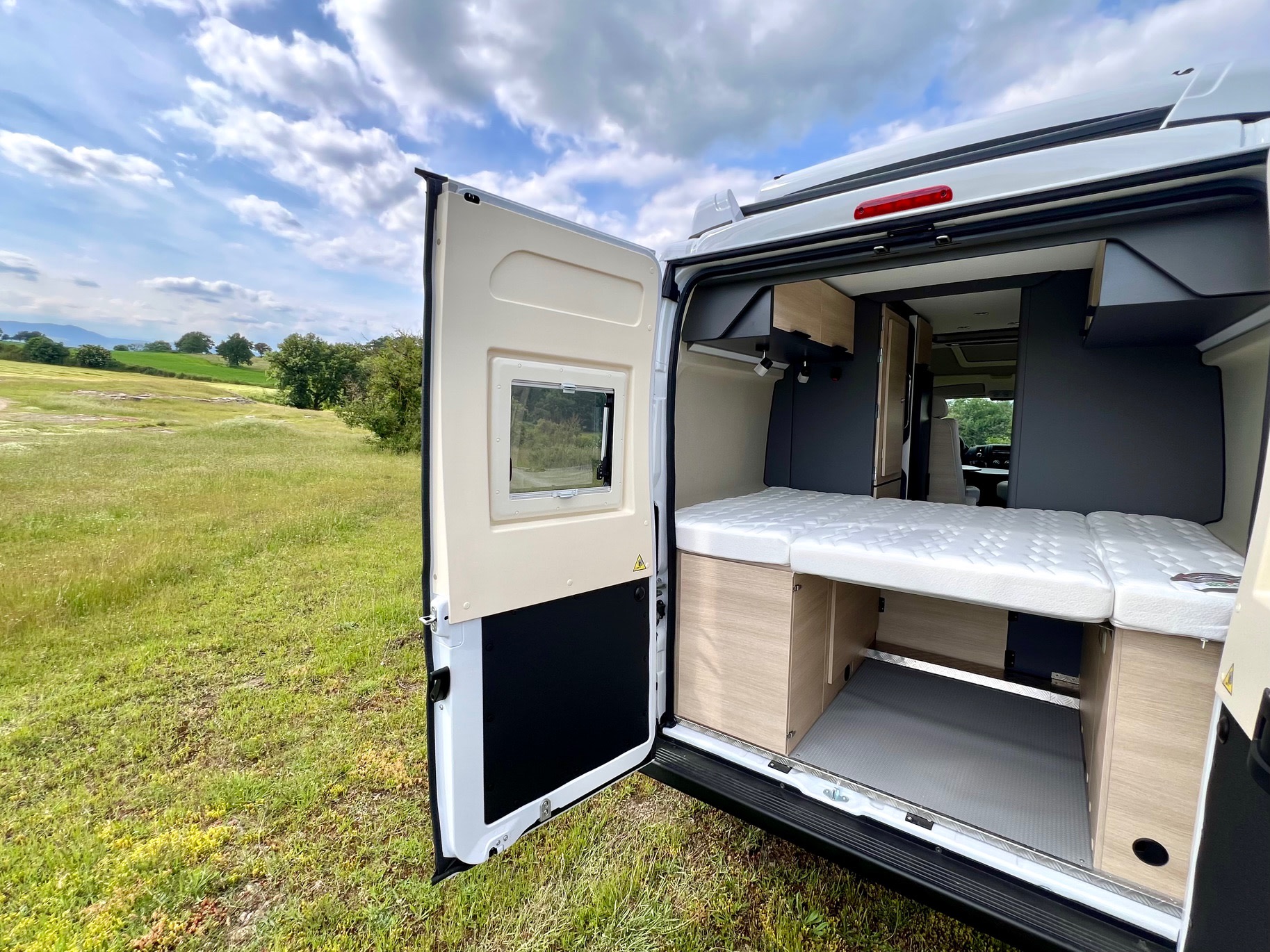 Inside a modern rv rental: a comfy bed awaits, framed by an open door and a lush green field view.. Peugeot Partner 2025