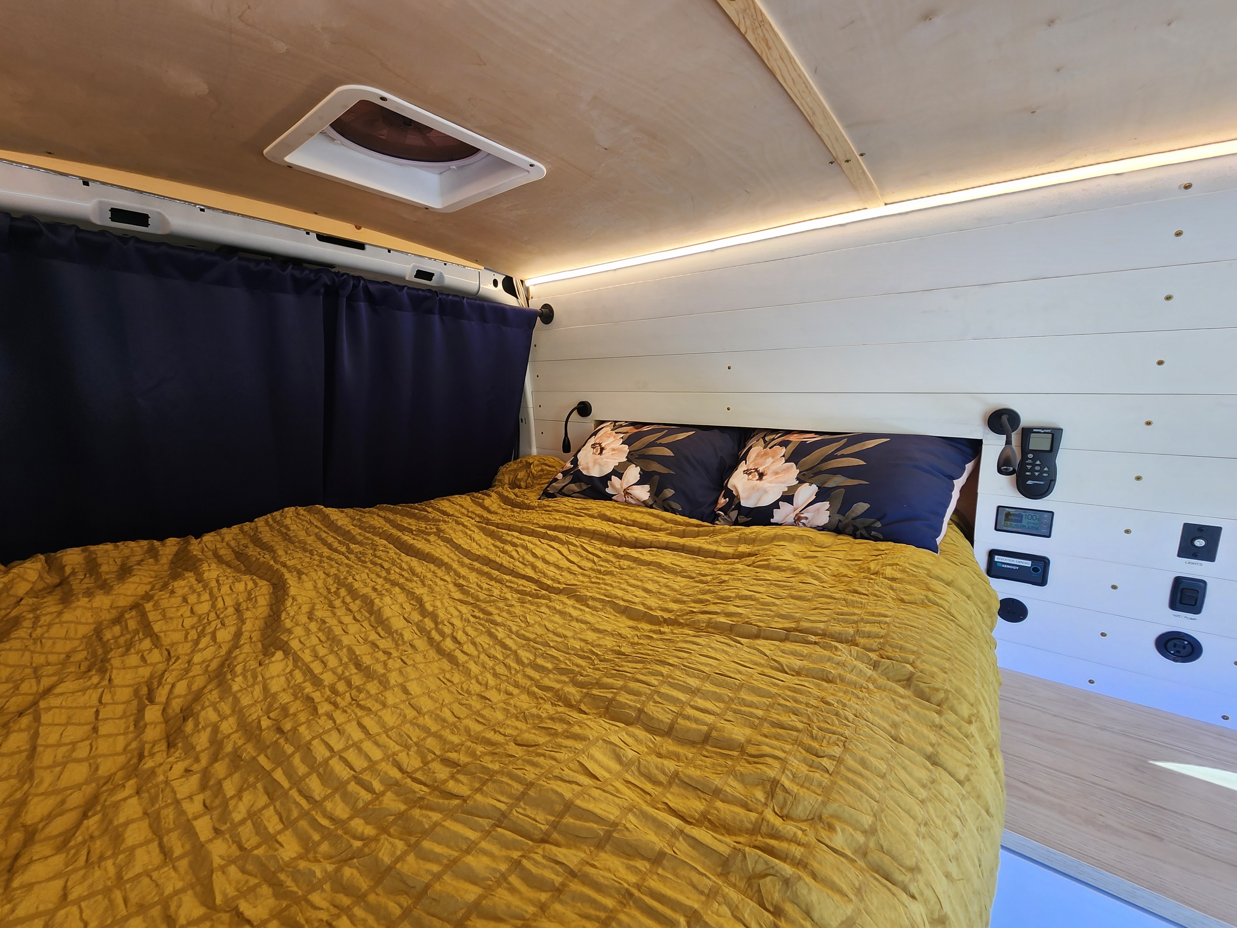 Inside a comfortable RV rental: A bed with a yellow comforter, floral pillows, and wall-mounted controls.. Ford Transit 2022