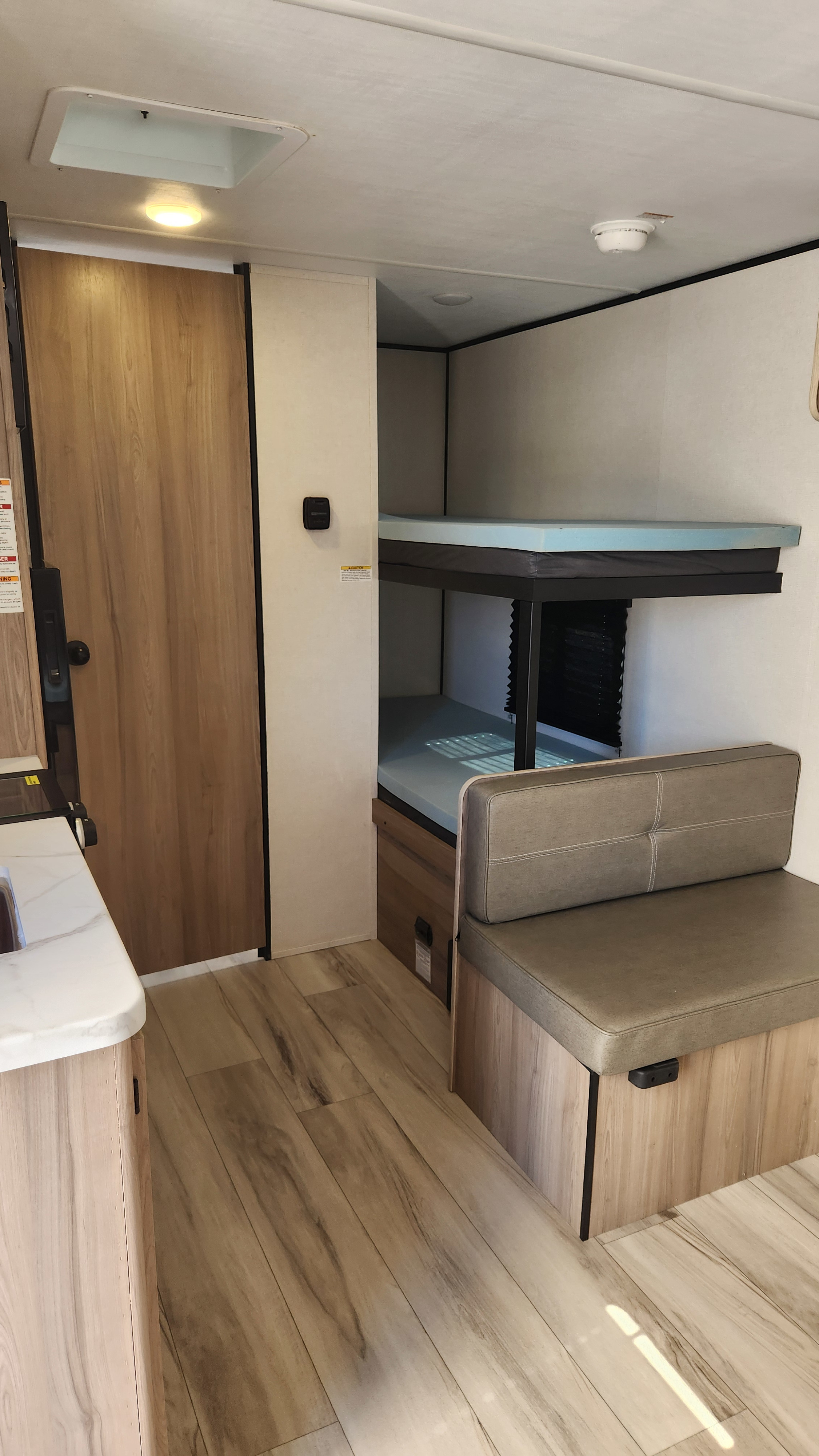 Bright RV rental interior with light wood floor, compact kitchen, bunk beds with blue mattresses, and gray dinette seating.. Keystone Coleman Light 17BX 2025