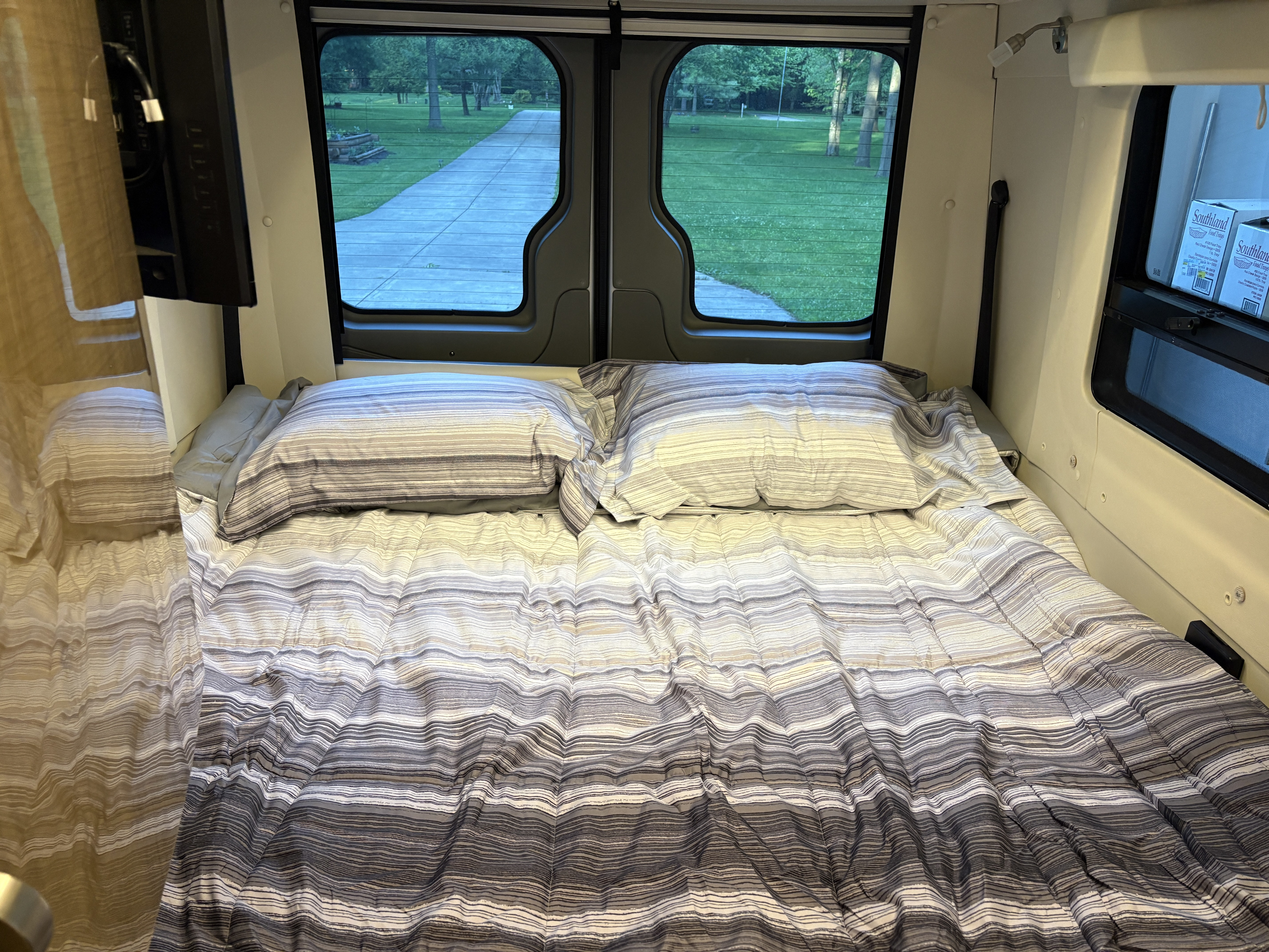 Cozy RV rental bed with striped bedding and a view of a park-like setting from the rear windows.. Winnebago Era 2017