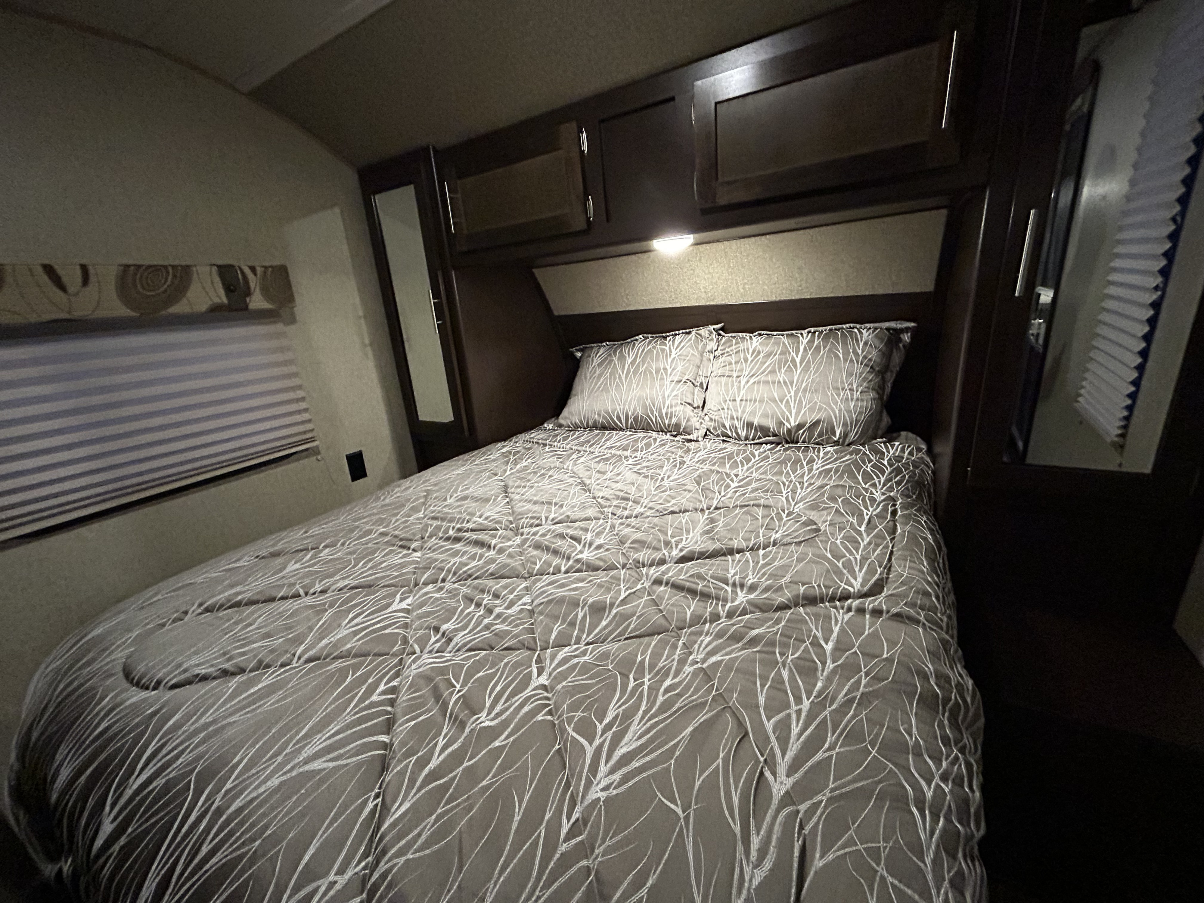 Cozy RV rental bedroom featuring a patterned bedspread, overhead cabinets, a window with blinds, and soft lighting.. Forest River Cherokee 2016
