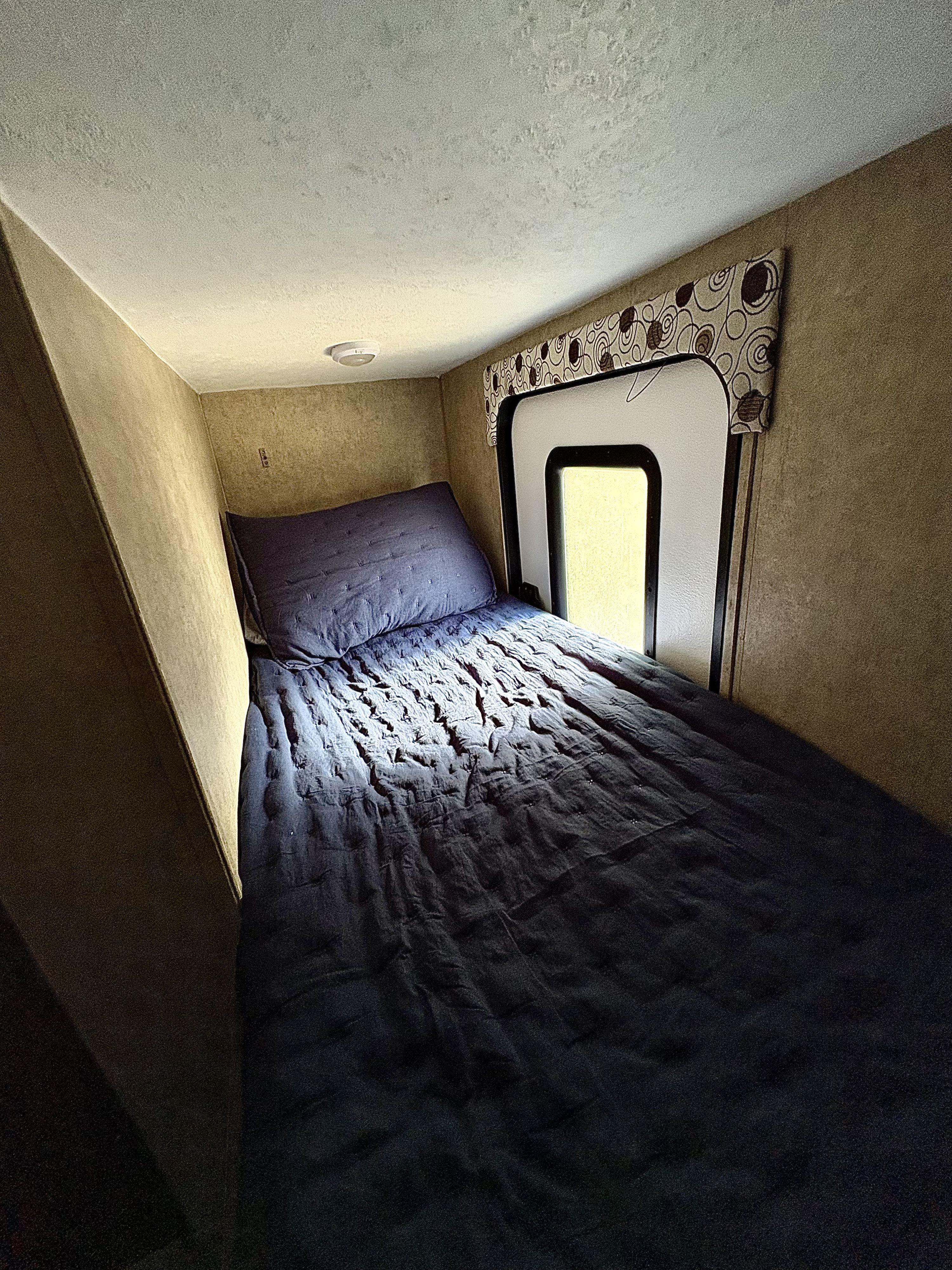 Small, cozy RV rental bedroom features a dark blue bed, pillow, and window with a patterned valance.. Coachmen Clipper 2017