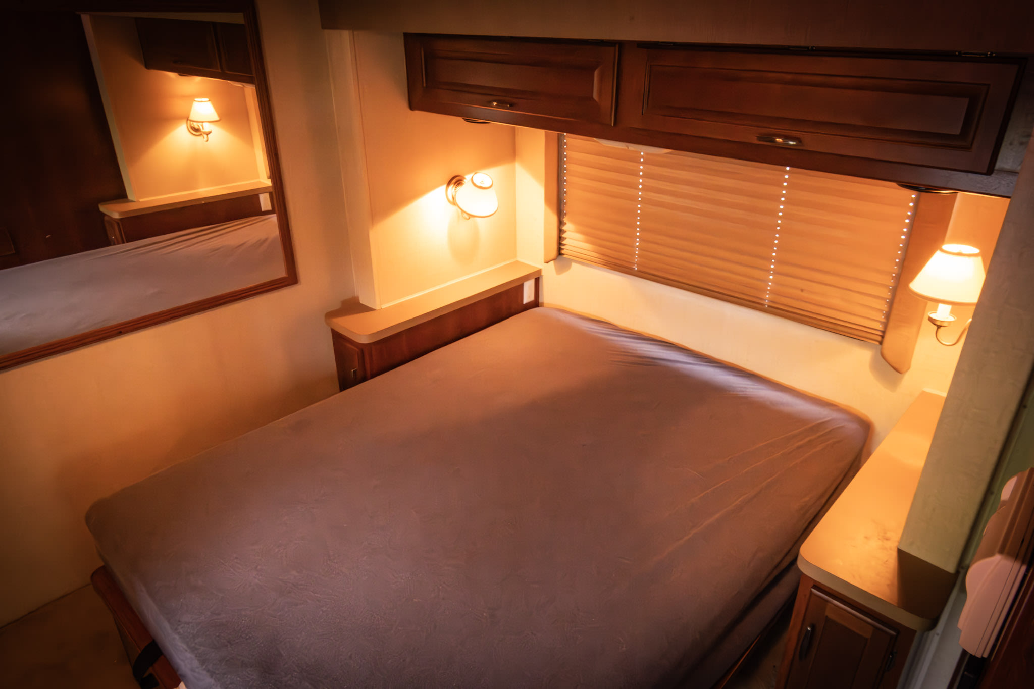 RV rental bedroom with a bed, warm sconce lighting, wood cabinets, blinds, and mirror.. Bounder Bounder Motorhome 2005