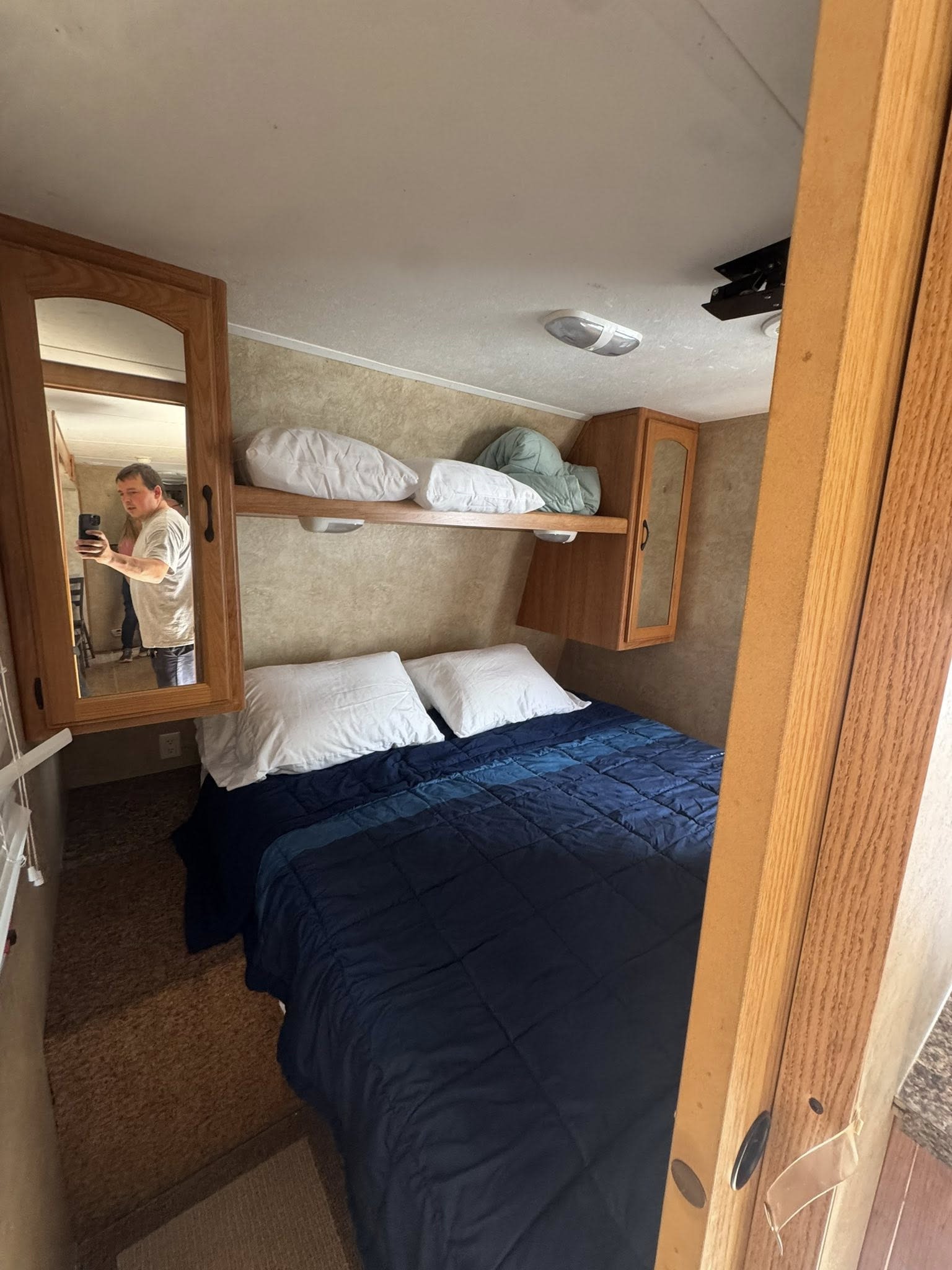 Cozy RV rental bedroom featuring a blue bed, white pillows, overhead cabinets, and a mirror reflecting a person.. Keystone RV Sprinter Select 2010