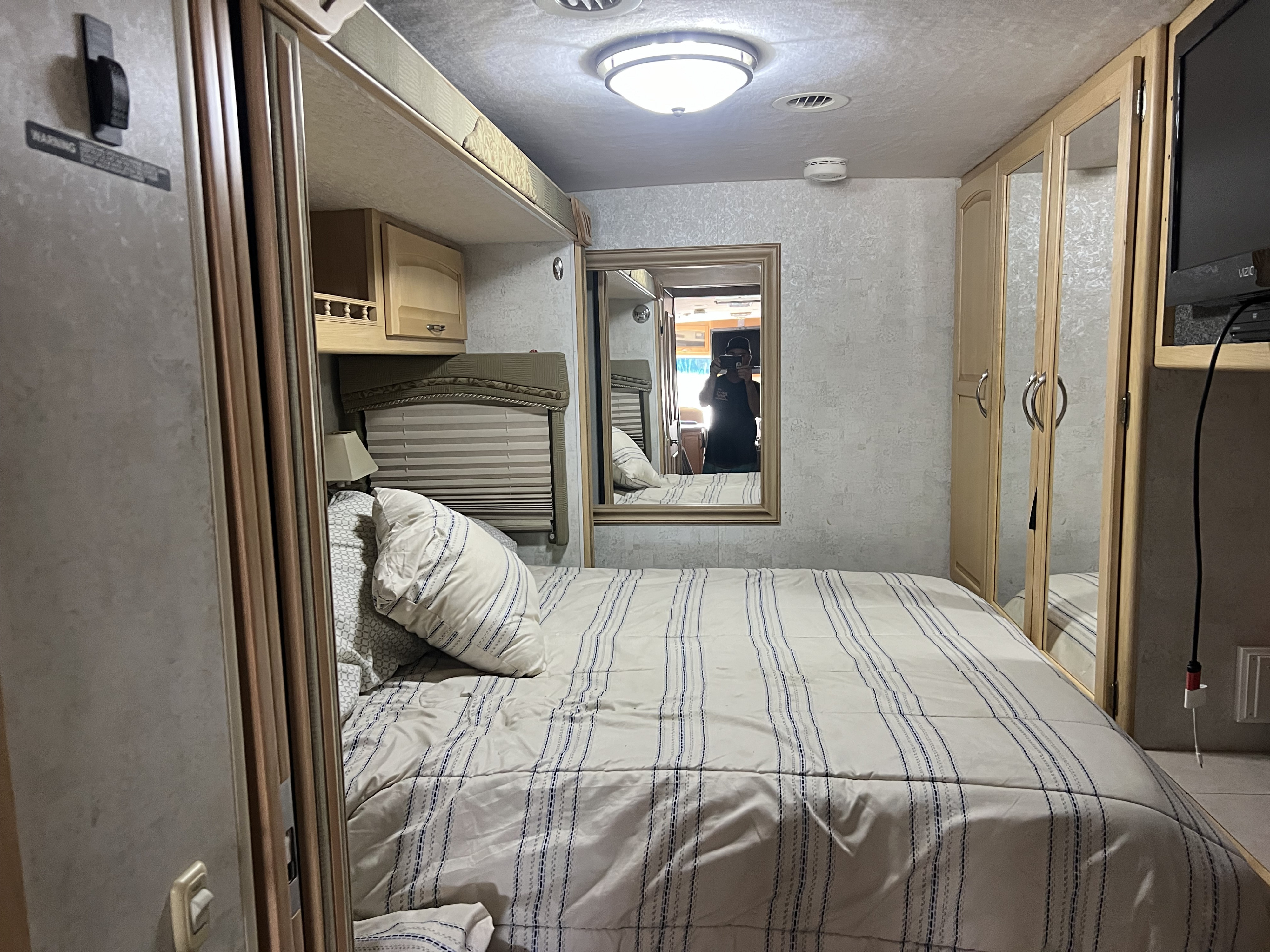 This cozy rv rental bedroom showcases a striped bed, overhead storage, a lamp, and a mirror reflecting the photographer.. Winnebago Ifg33v 2005