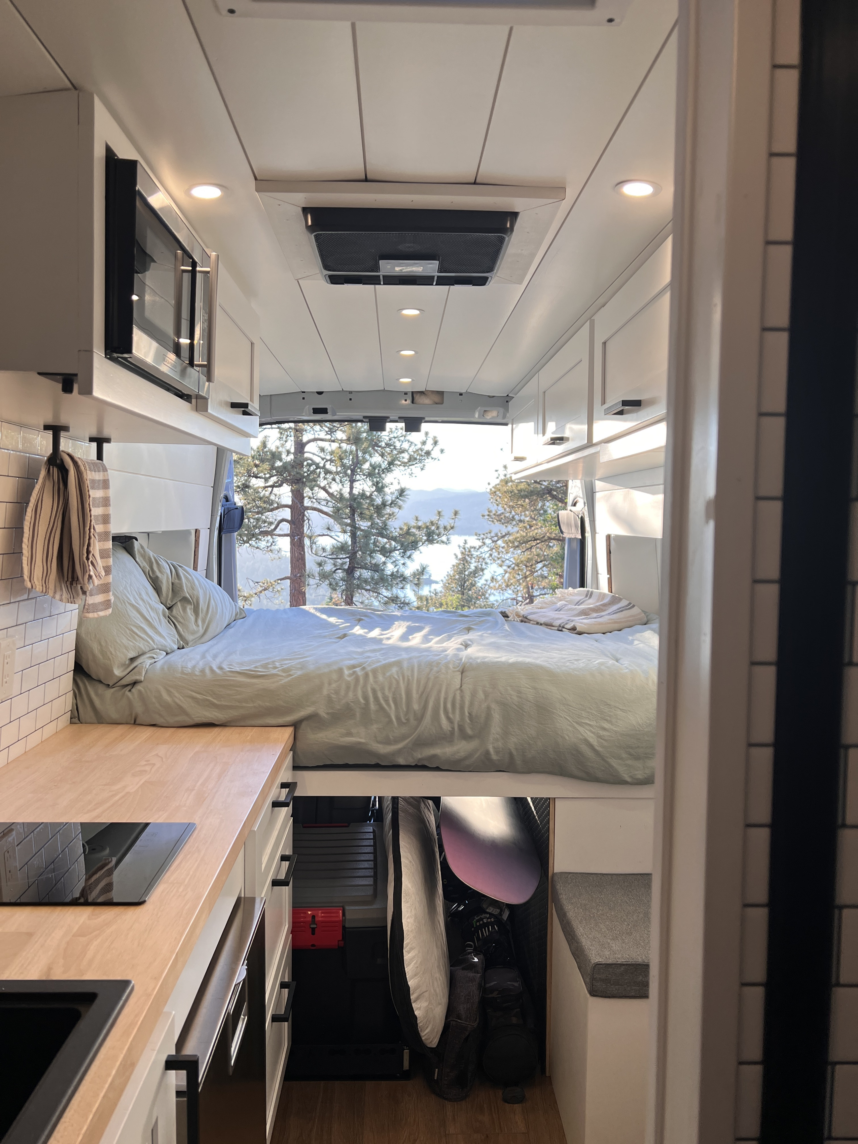 Cozy rv rental interior: van bed overlooks a scenic lake & pine trees, with a compact kitchen in foreground.. ActiVan Ford Transit 350 AWD 2023