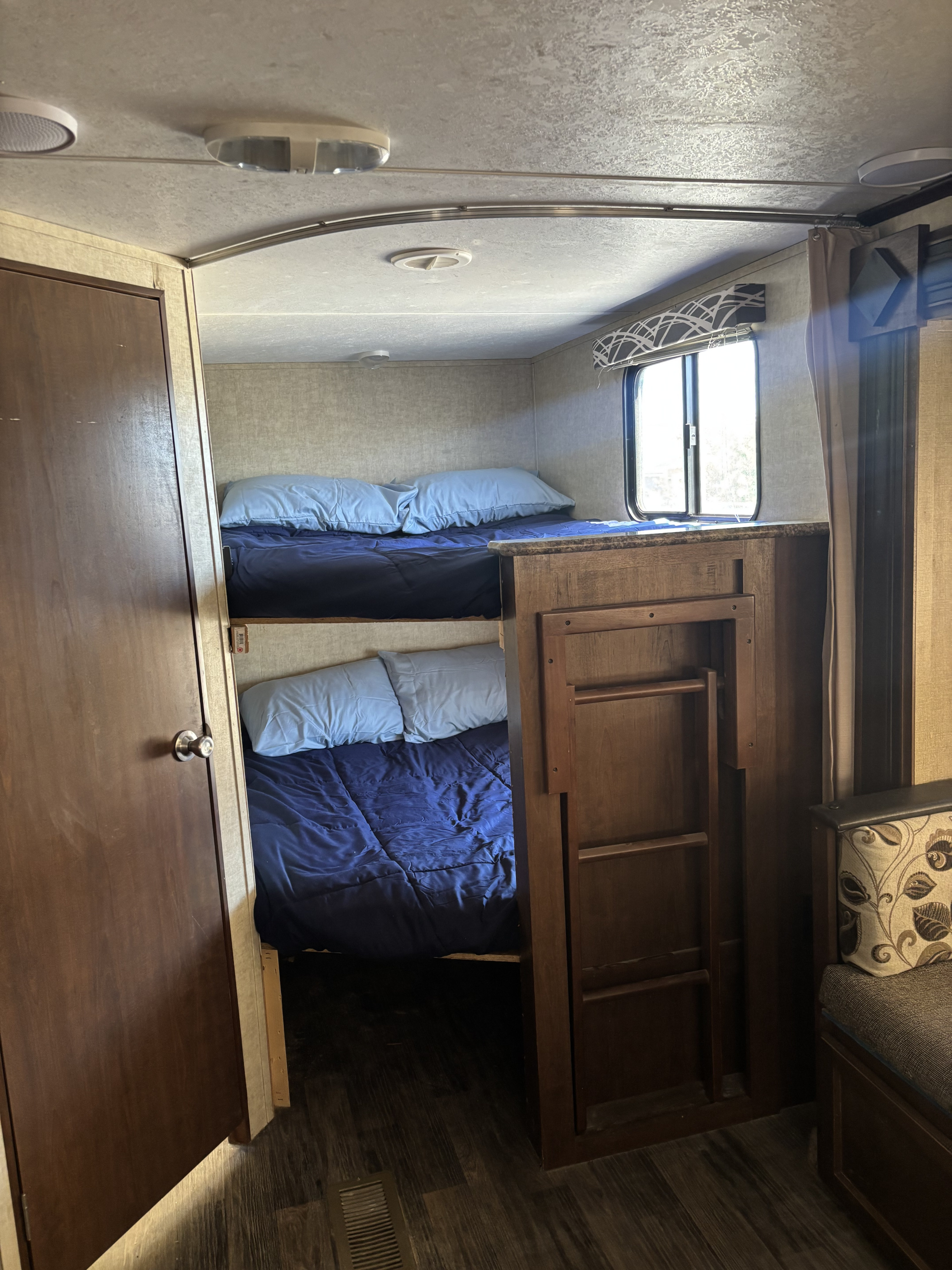 Inside an RV rental, showing a comfortable bunk bed sleeping area with blue linens, an access ladder, and a window.. Keystone RV 28BHS 2016