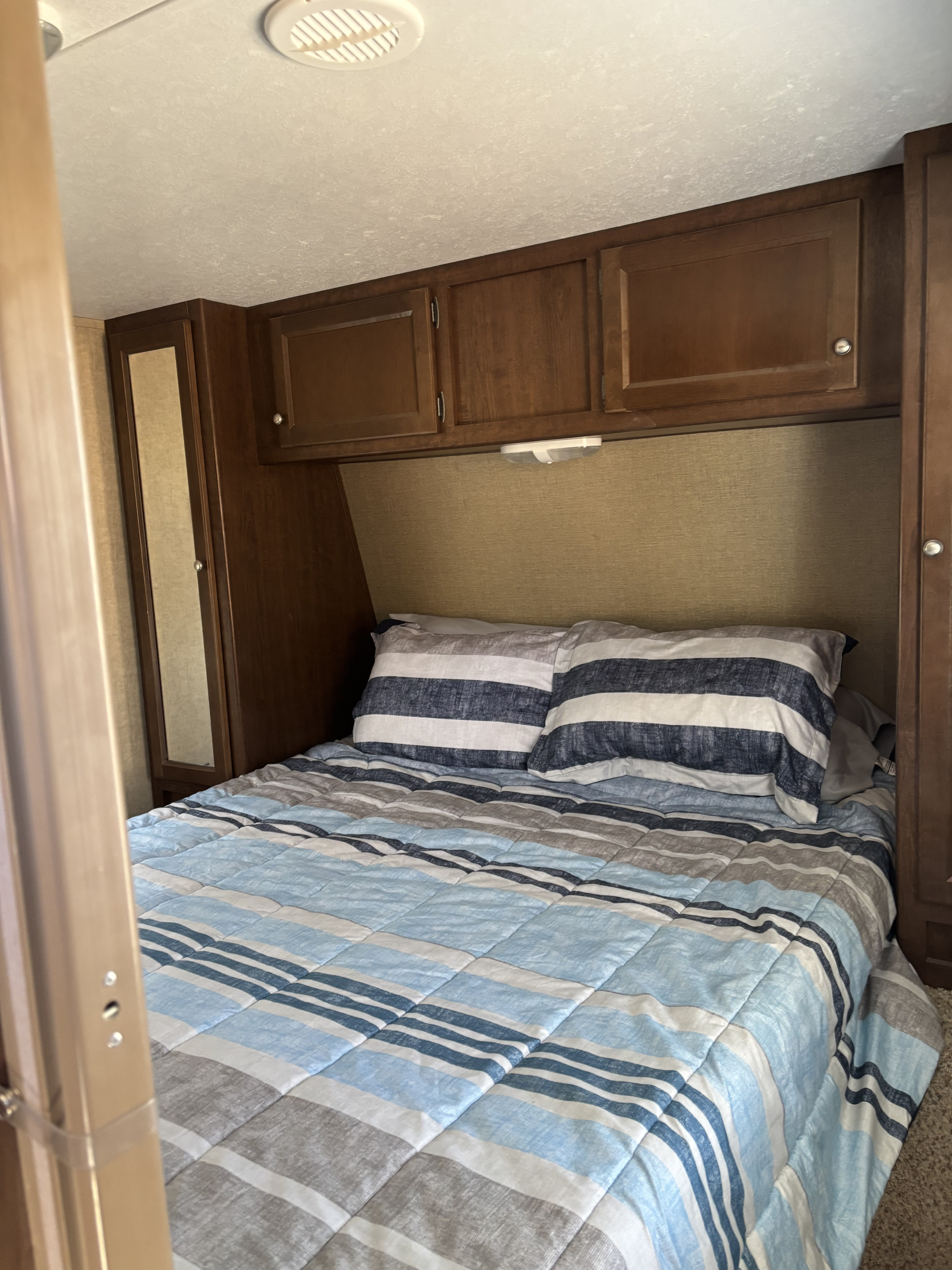 Inside a cozy rv rental bedroom: a bed with striped blue, white, and gray bedding, overhead wooden cabinets, and mirrored storage.. Keystone RV 28BHS 2016
