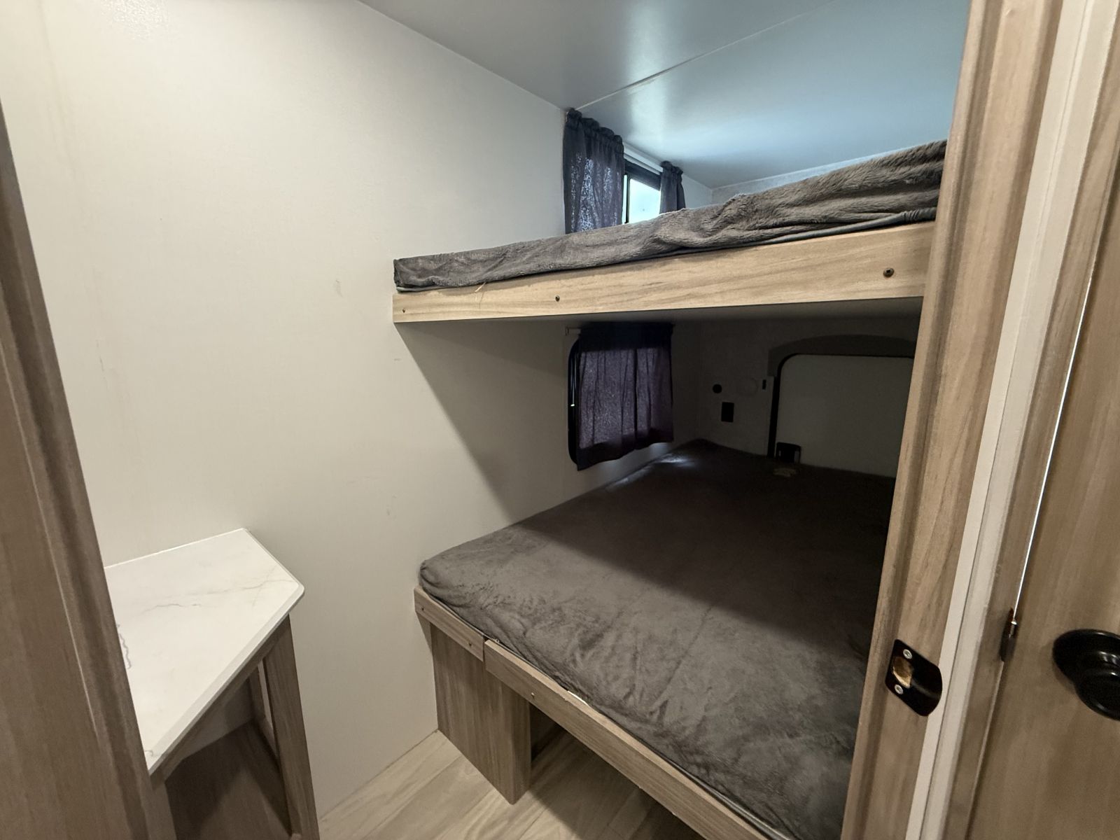 Cozy RV rental bunk beds featuring light wood frames, dark grey bedding, and a small white table in a compact sleeping area.. Winnebago Access 2024
