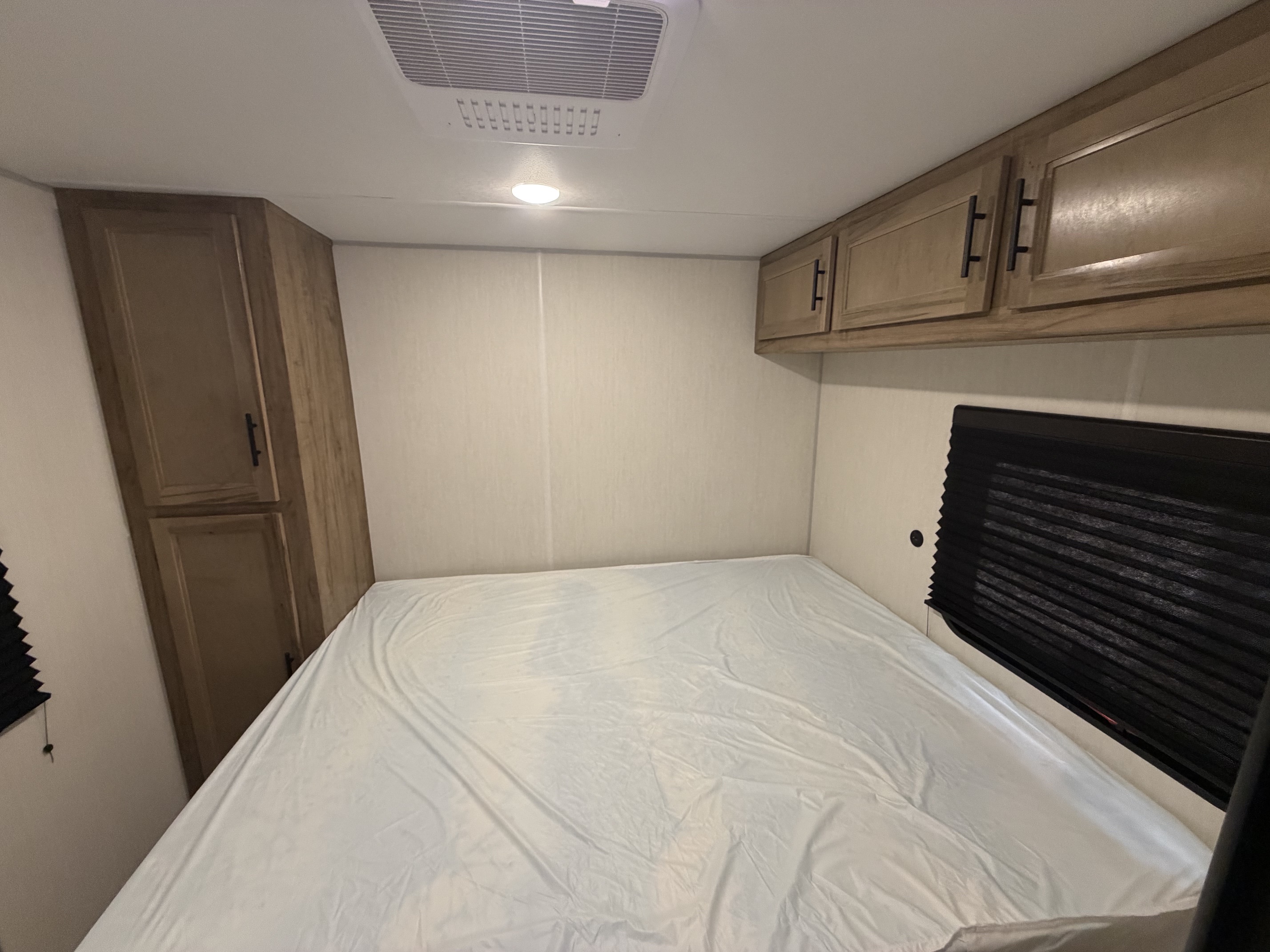 Cozy RV rental bedroom features a bed with white sheets, wood cabinets, AC, and a window with a blind.. Forest River Cherokee Grey Wolf 2025