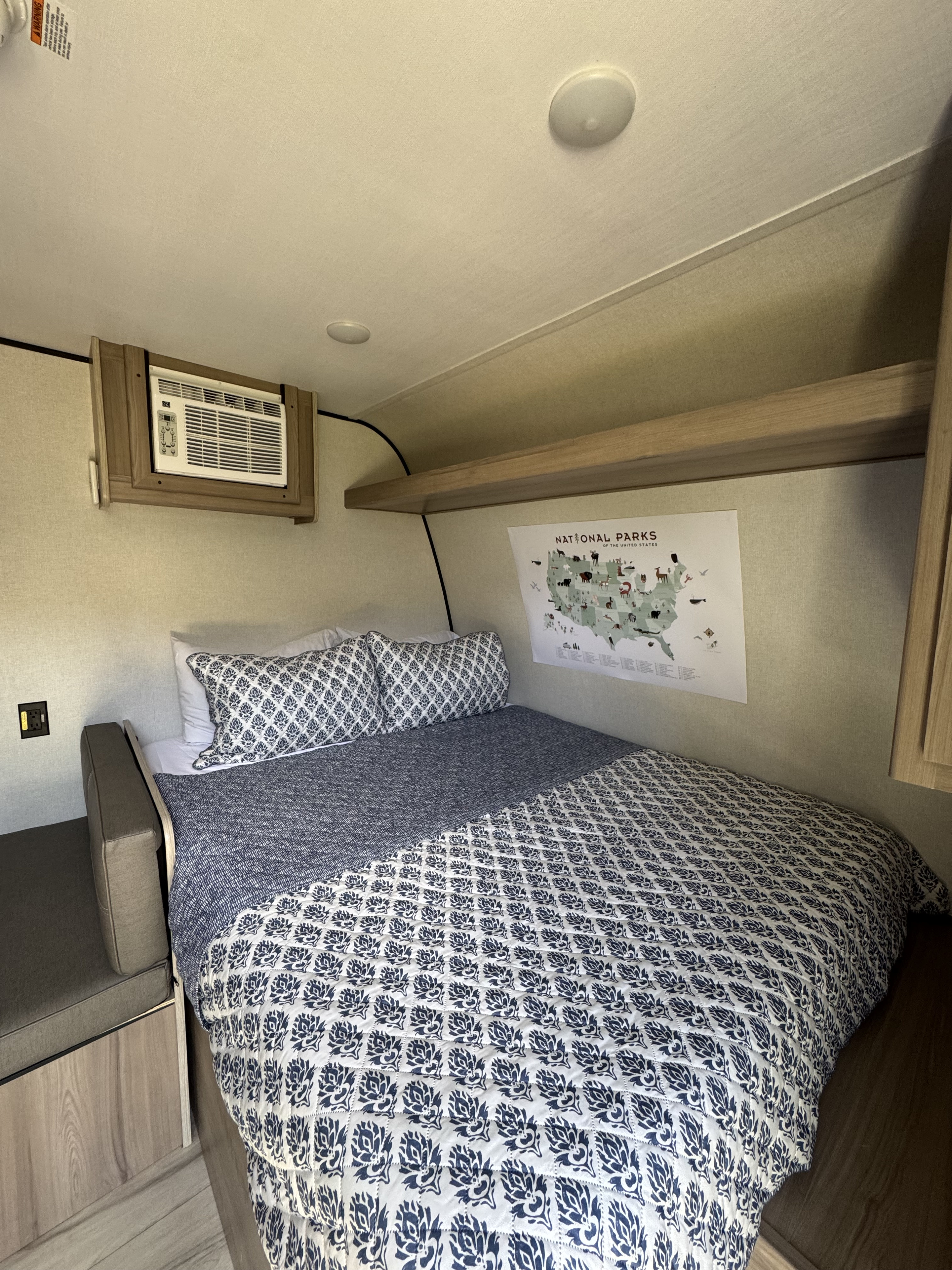 This RV rental bedroom features a cozy bed with patterned blue and white bedding, a wall AC unit, and a National Parks map.. Dutchmen Coleman 2025