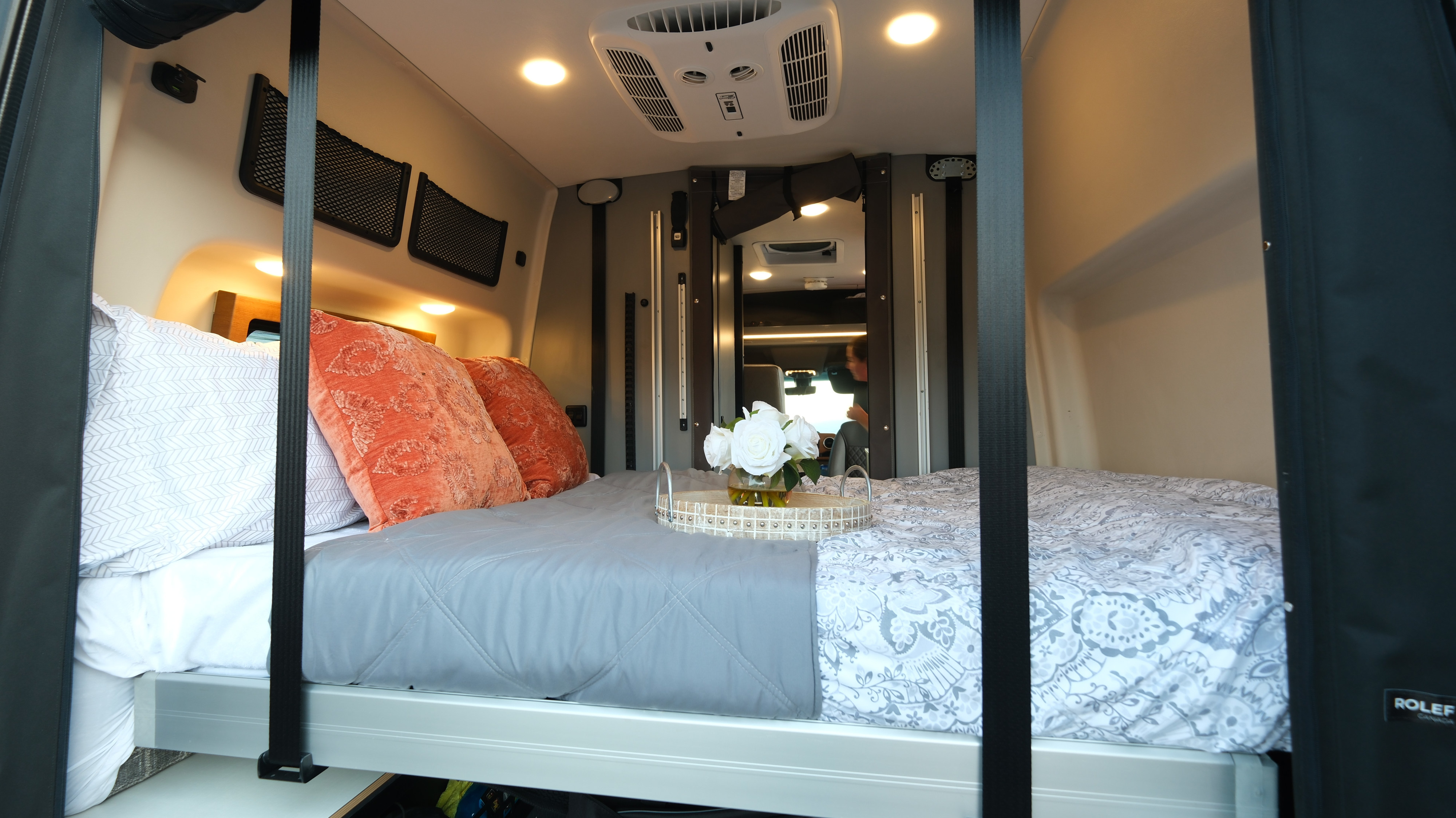 This RV rental interior features a cozy bed with orange pillows, overhead lights, storage nets, and a decorative tray with white roses.. Other Other 2026
