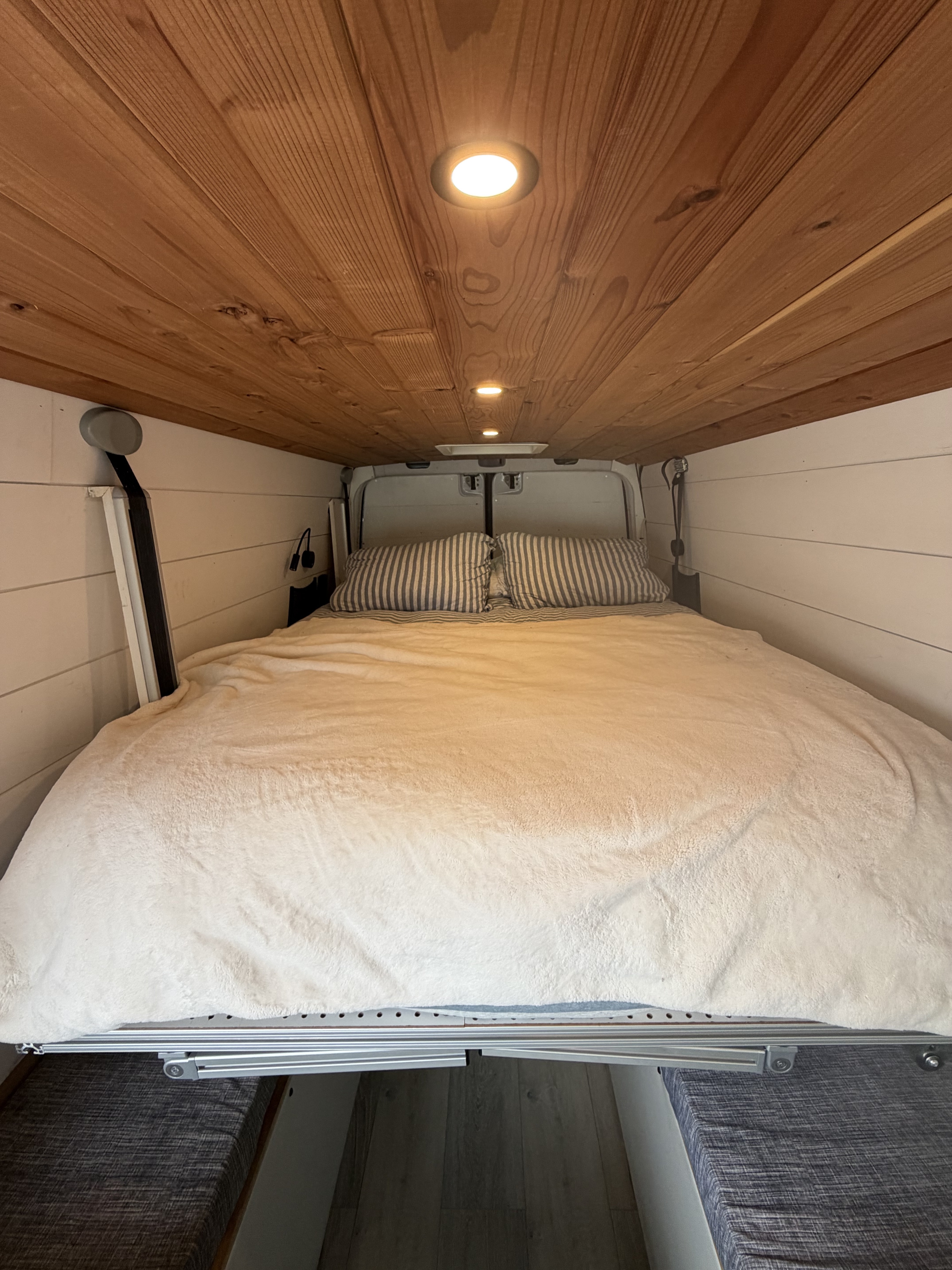Cozy RV rental interior shows a large bed with striped pillows, a wooden ceiling with lights, and white paneled walls.. Mercedes Sprinter 2010