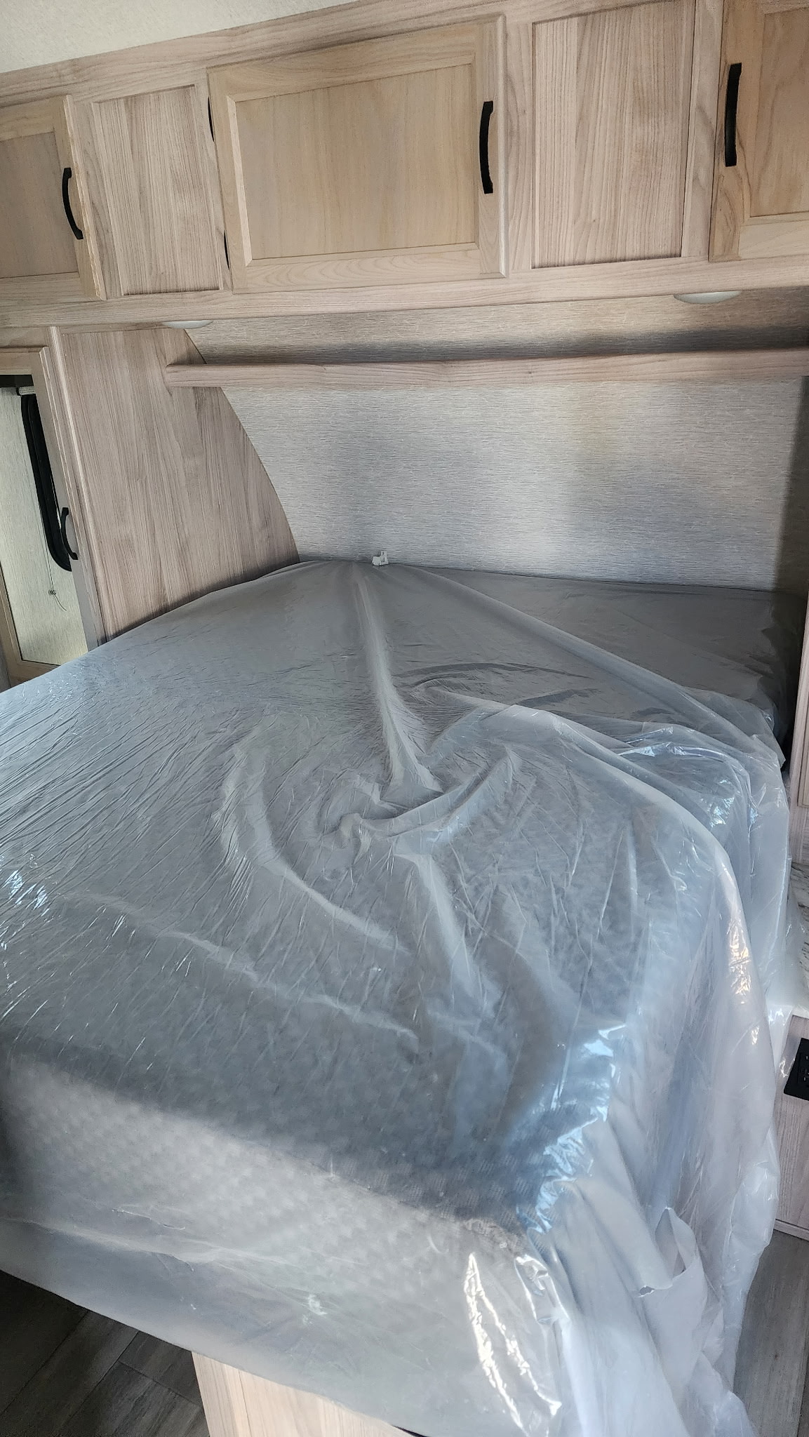New rv rental bedroom with a bed covered in protective plastic, overhead light wood cabinets, and shelf.. Coachmen 292bhs 2021