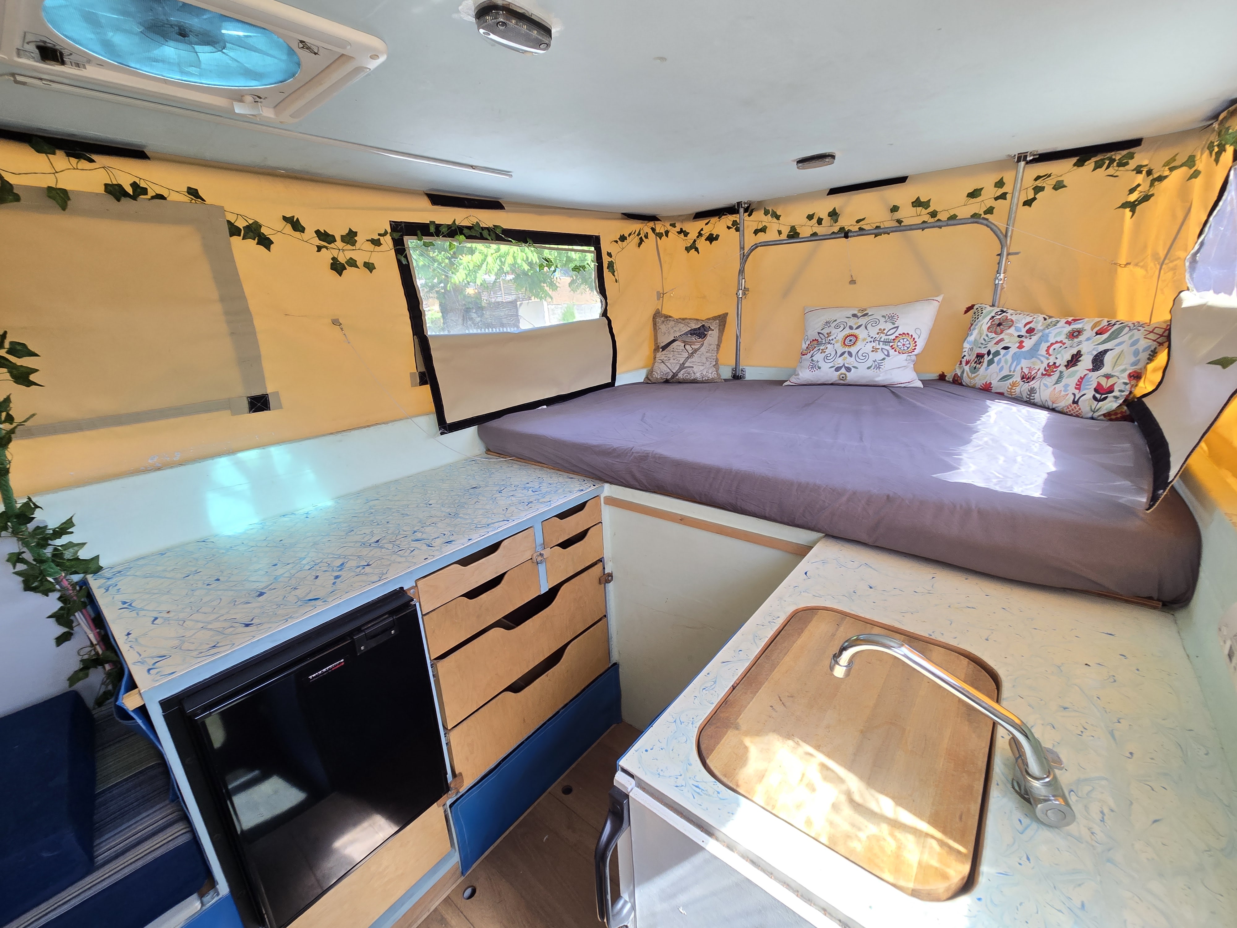 Cozy rv rental interior featuring a bed with pillows, kitchen counter with sink, fridge, drawers, and decorative ivy.. Toyota Tundra 2005