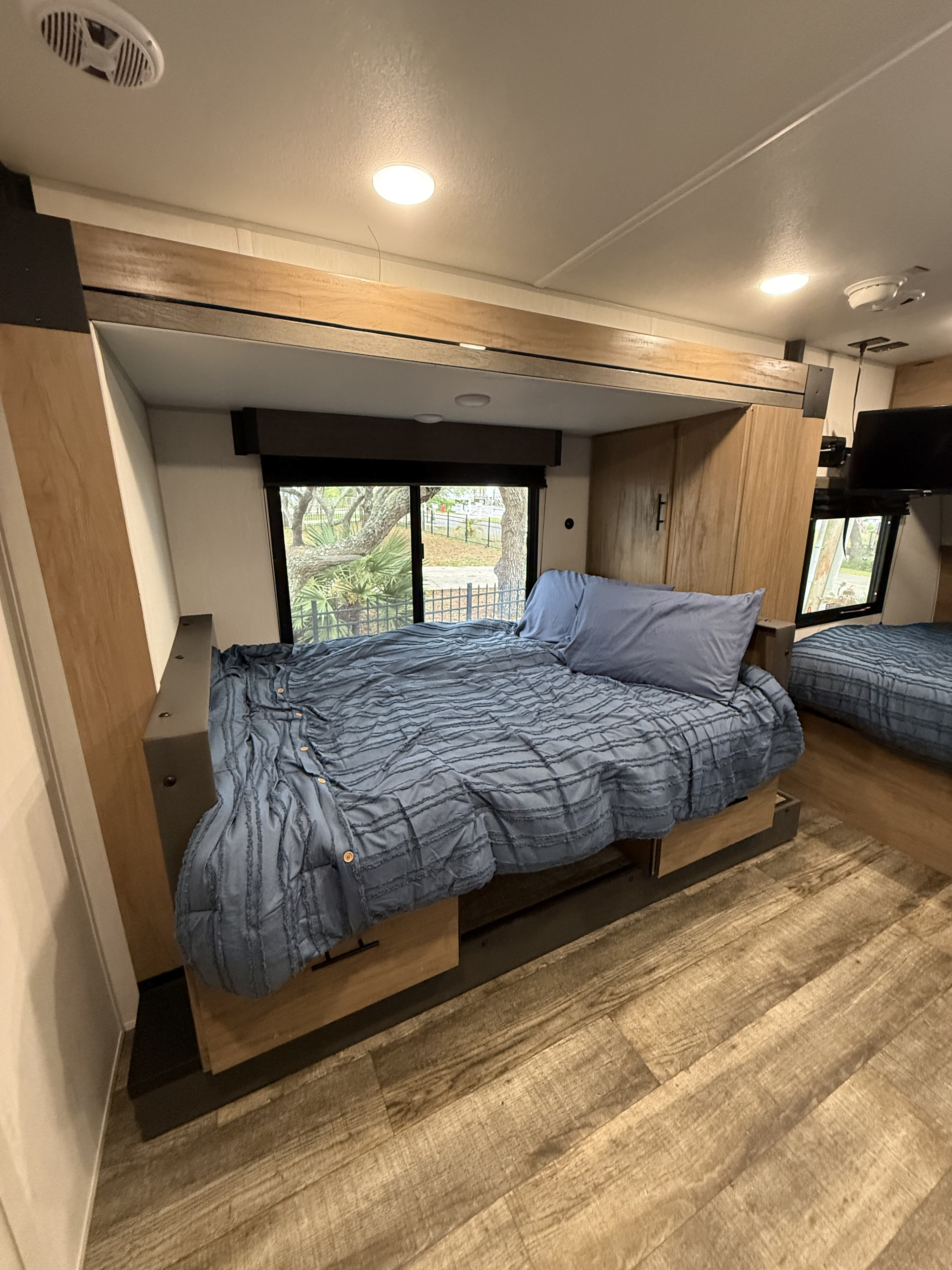 Modern RV rental interior: bedroom featuring blue beds with plush bedding, wood flooring, and a window overlooking trees.. Forest River Campsite Reserve LS17 2025