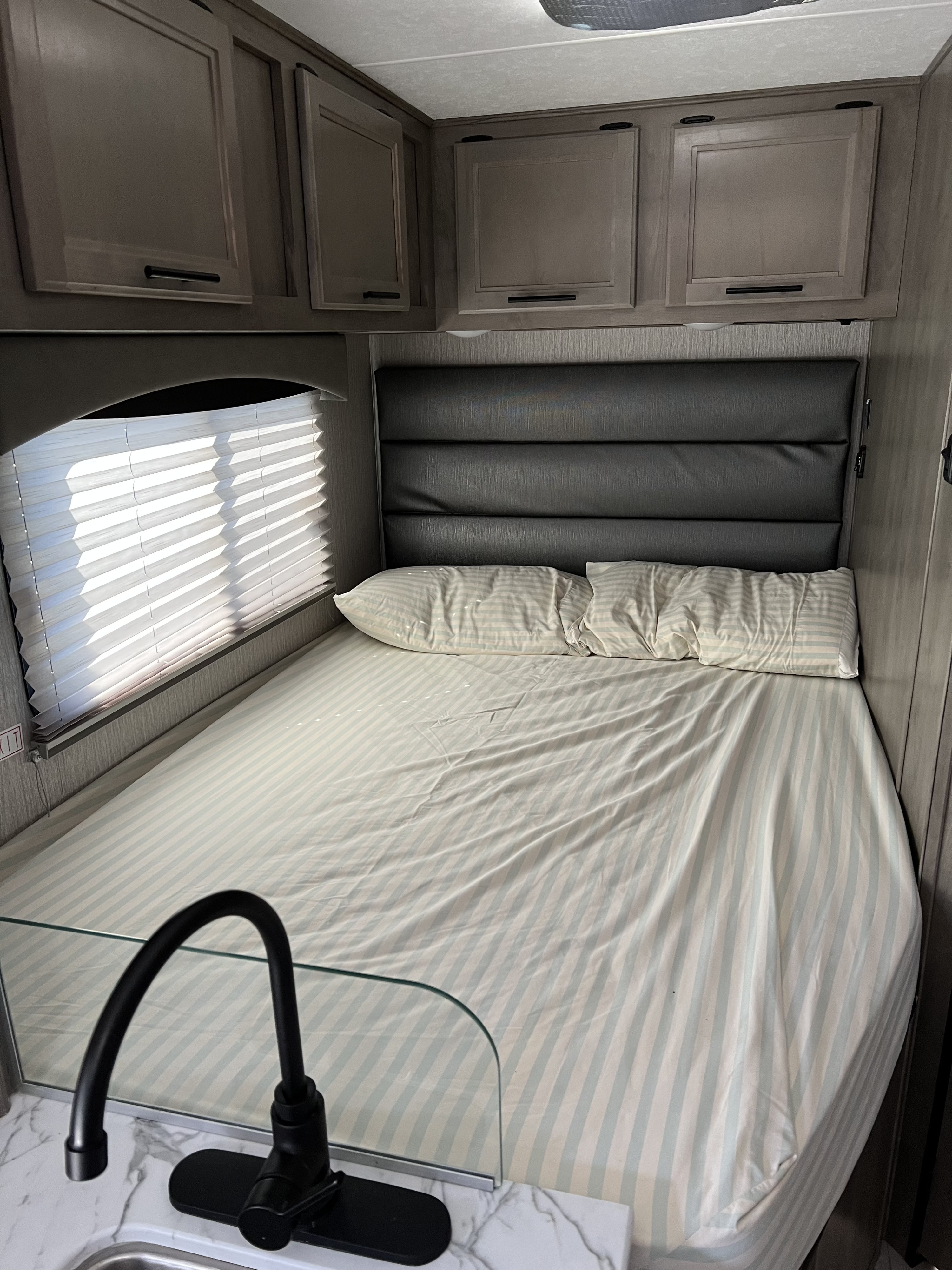 Cozy rv rental bedroom area with a striped bed, padded headboard, overhead cabinets, and a modern black faucet.. Forest River Coachmen Cross Trail XL 2022
