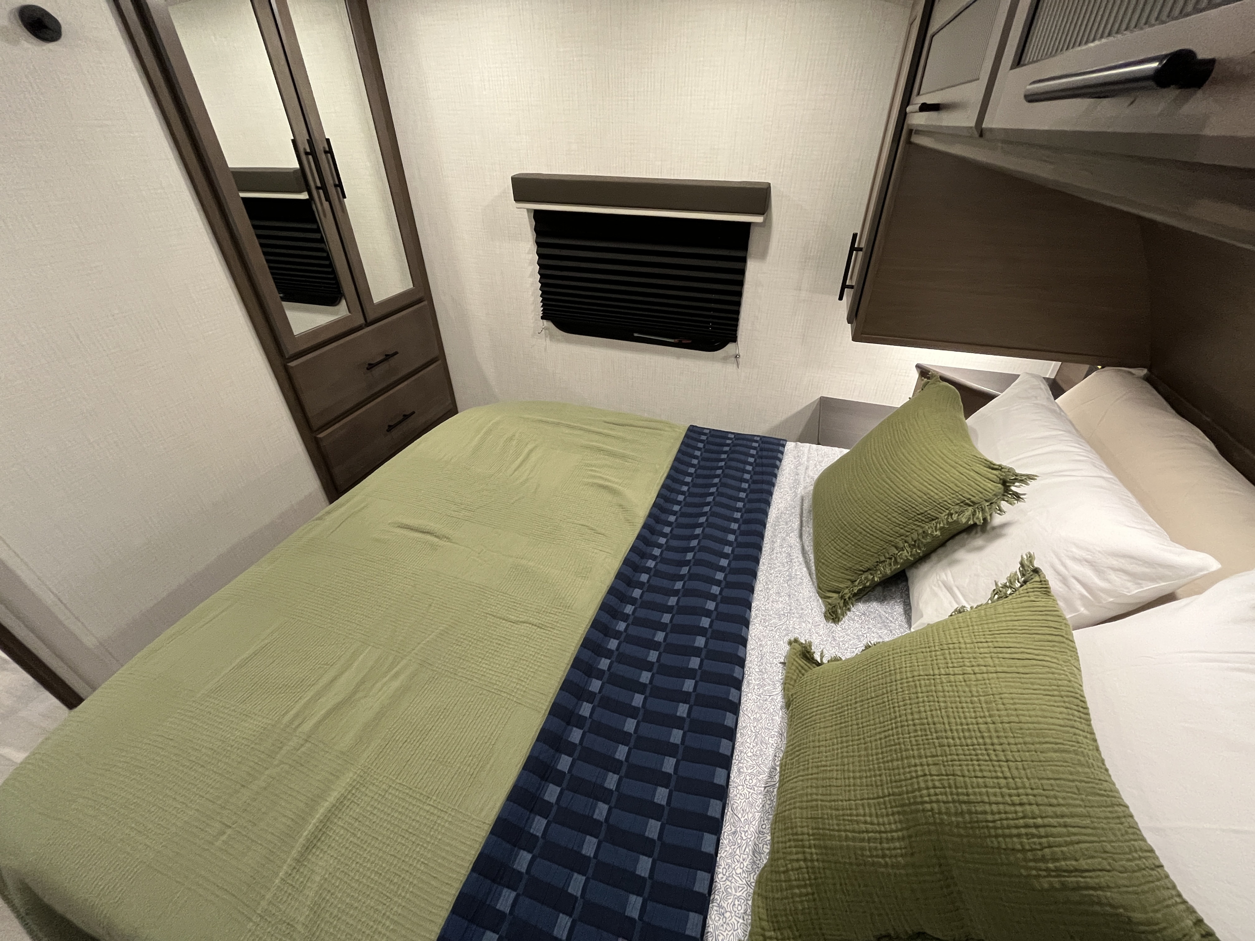 RV rental bedroom with a bed featuring green and blue bedding, mirrored wardrobe, overhead storage, and window blind.. Keystone RV Cougar 2026