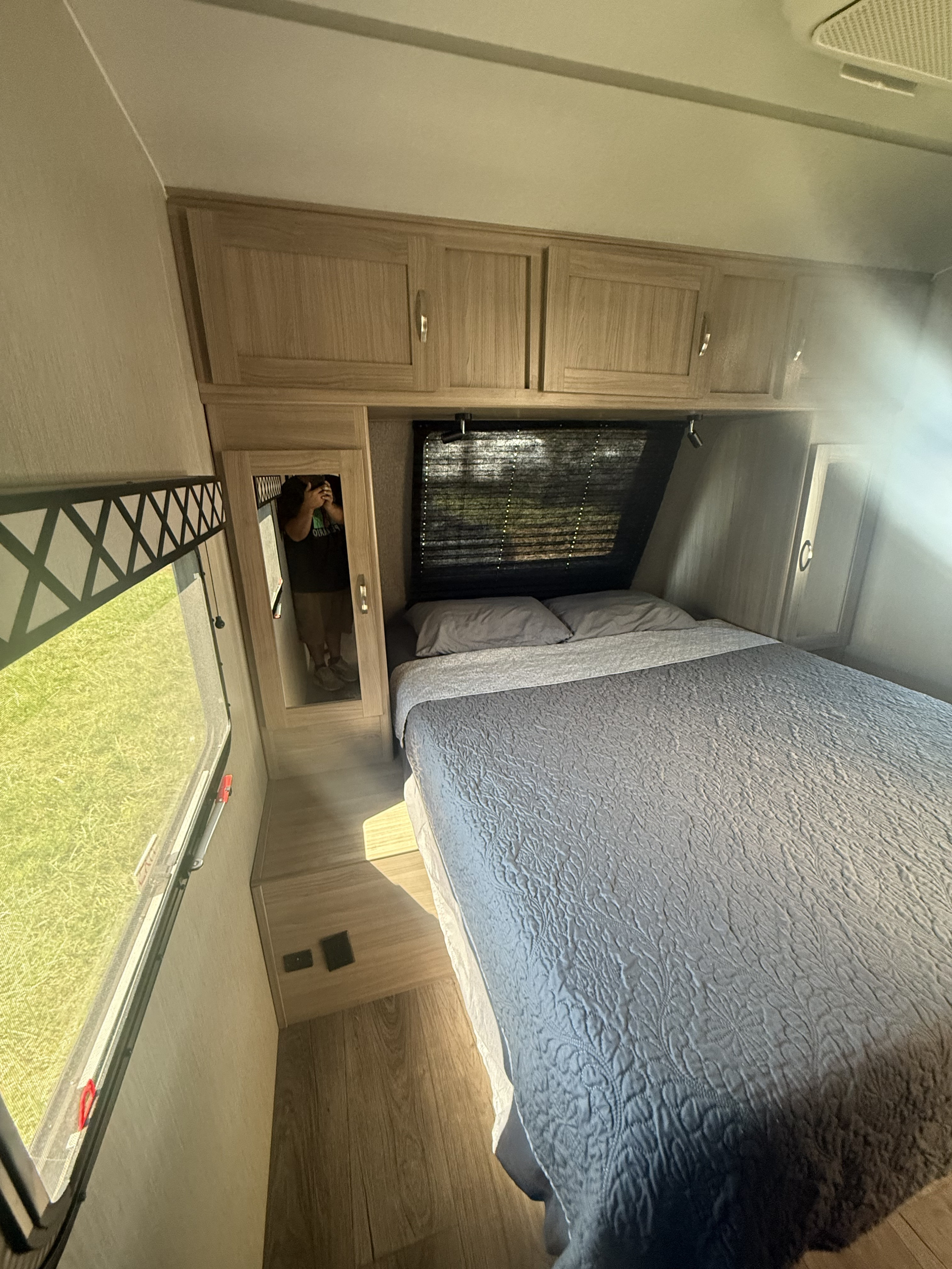 RV rental bedroom: gray bed, wood cabinets, window showing grass. Photographer visible in mirror.. Coachmen Other 2021